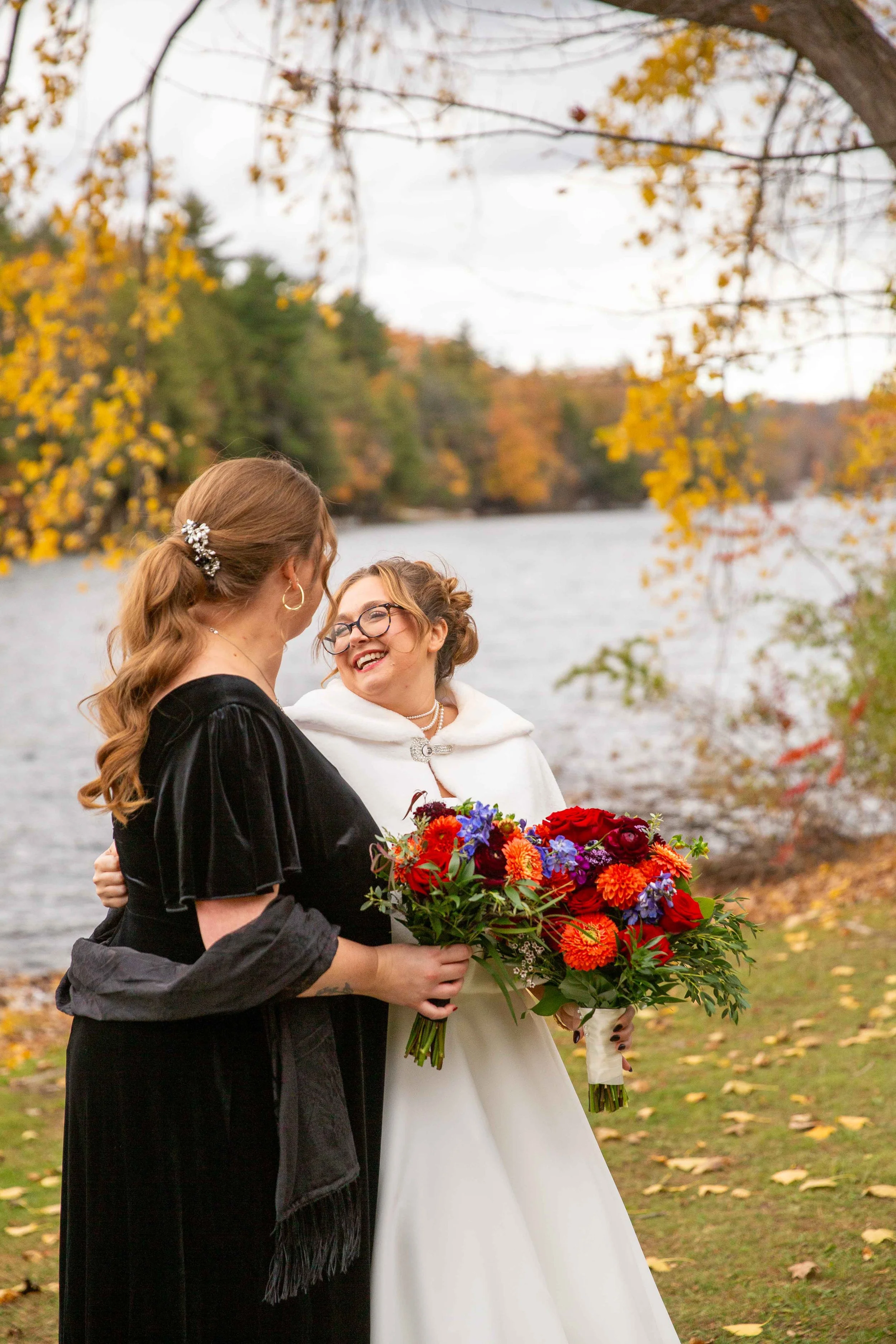 Two women smiling and enjoying a moment by a river with autumn trees. One woman is holding a bouquet of colorful flowers.