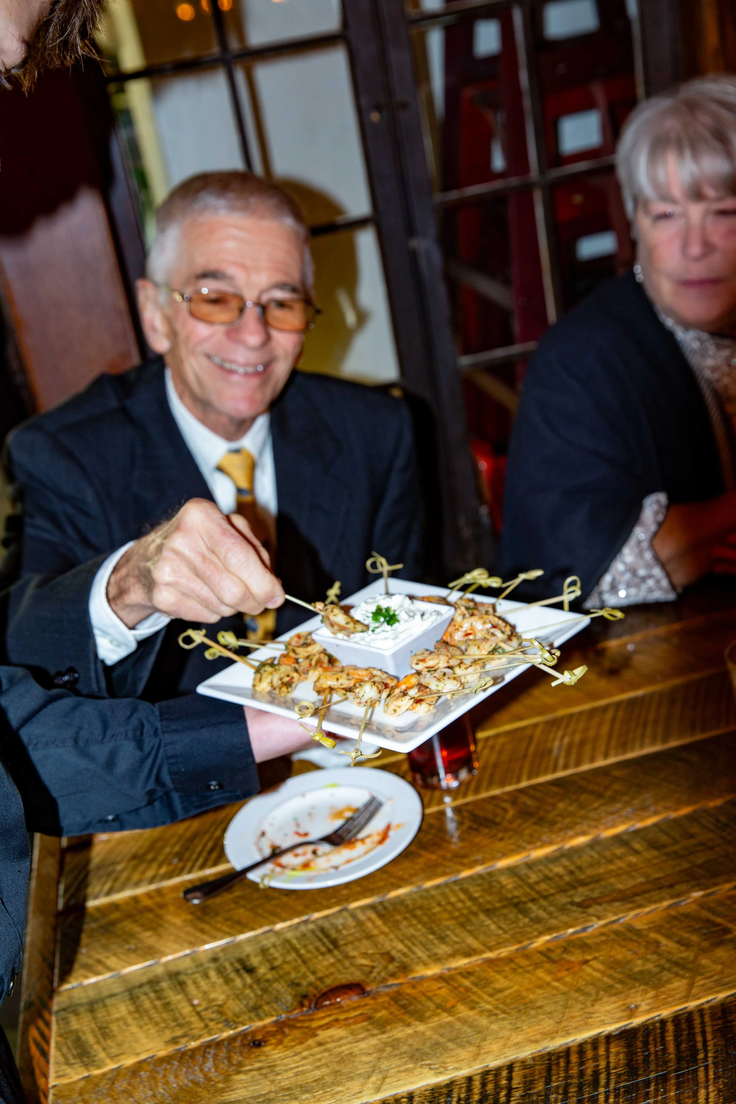 A man in a suit and glasses holding a tray of shrimp with a dipping sauce, sitting at a wooden table in a restaurant.