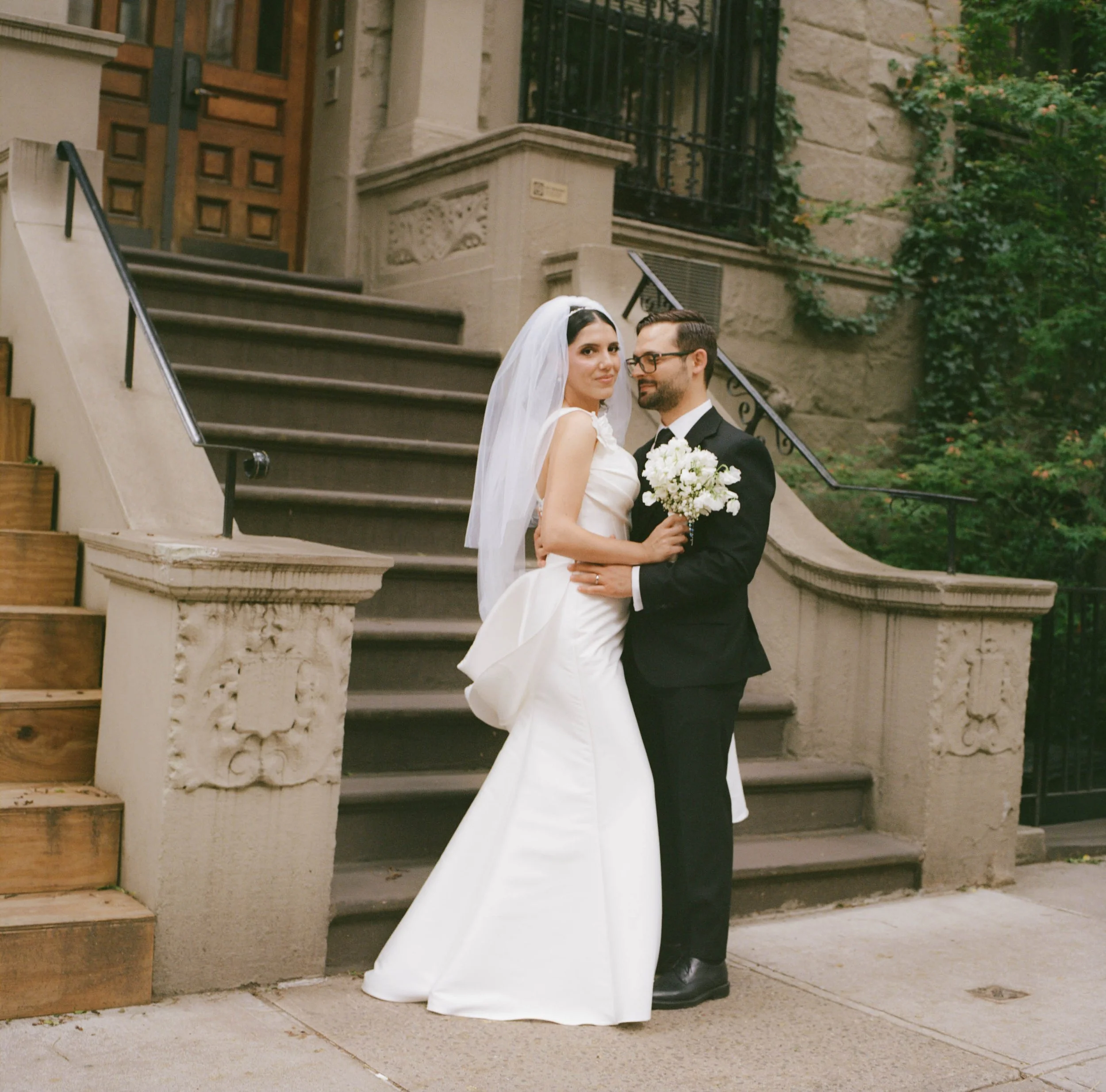 A bride and groom standing on a city sidewalk in front of stairs and a building. The bride is wearing a white wedding gown and veil, and the groom is dressed in a black suit and glasses. The bride is holding a bouquet of white flowers, and they are l