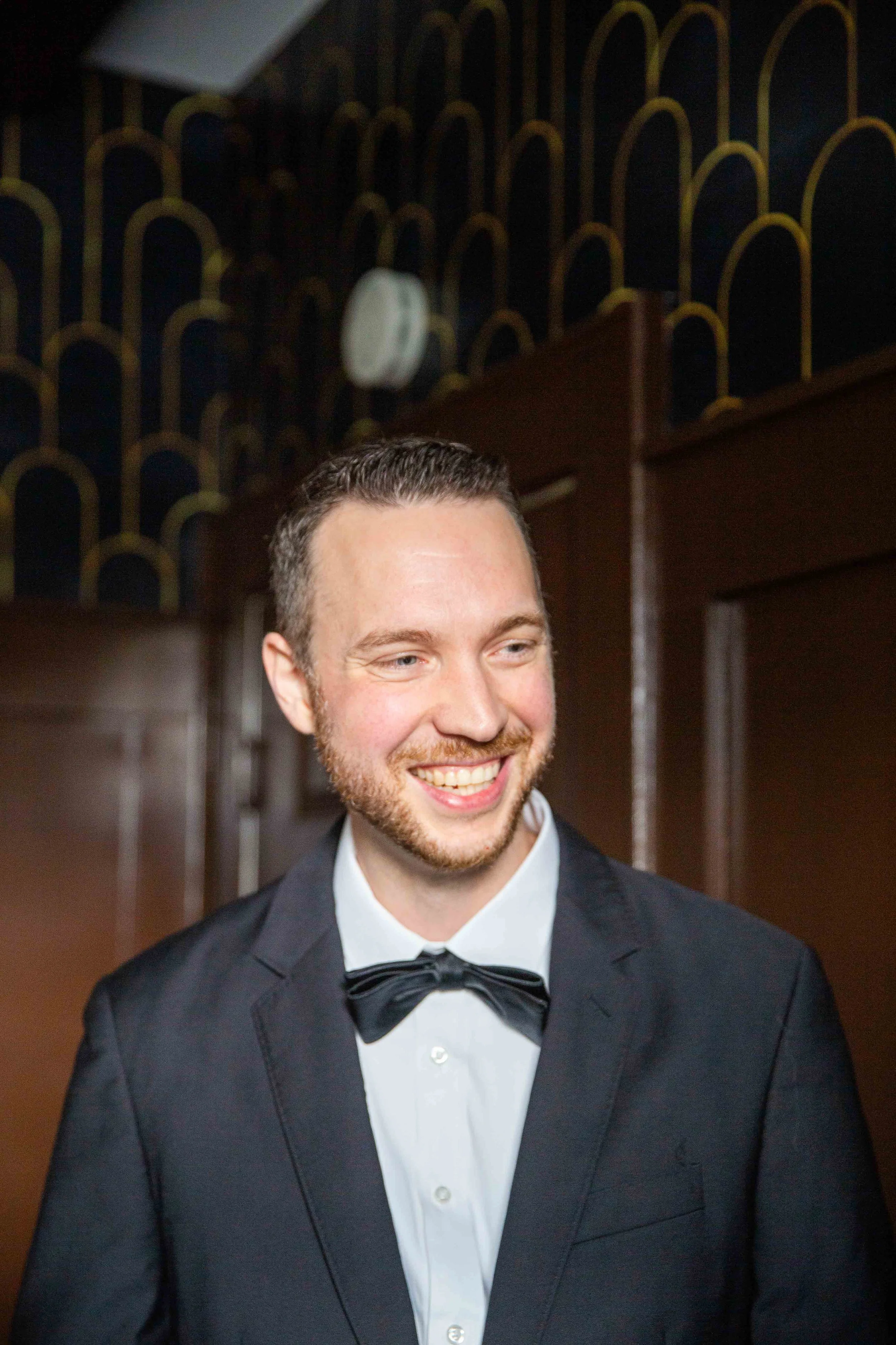 A young man with short dark hair and a beard, smiling, dressed in a black tuxedo with a white shirt and black bow tie, standing indoors with wooden paneled walls and patterned wallpaper in the background.