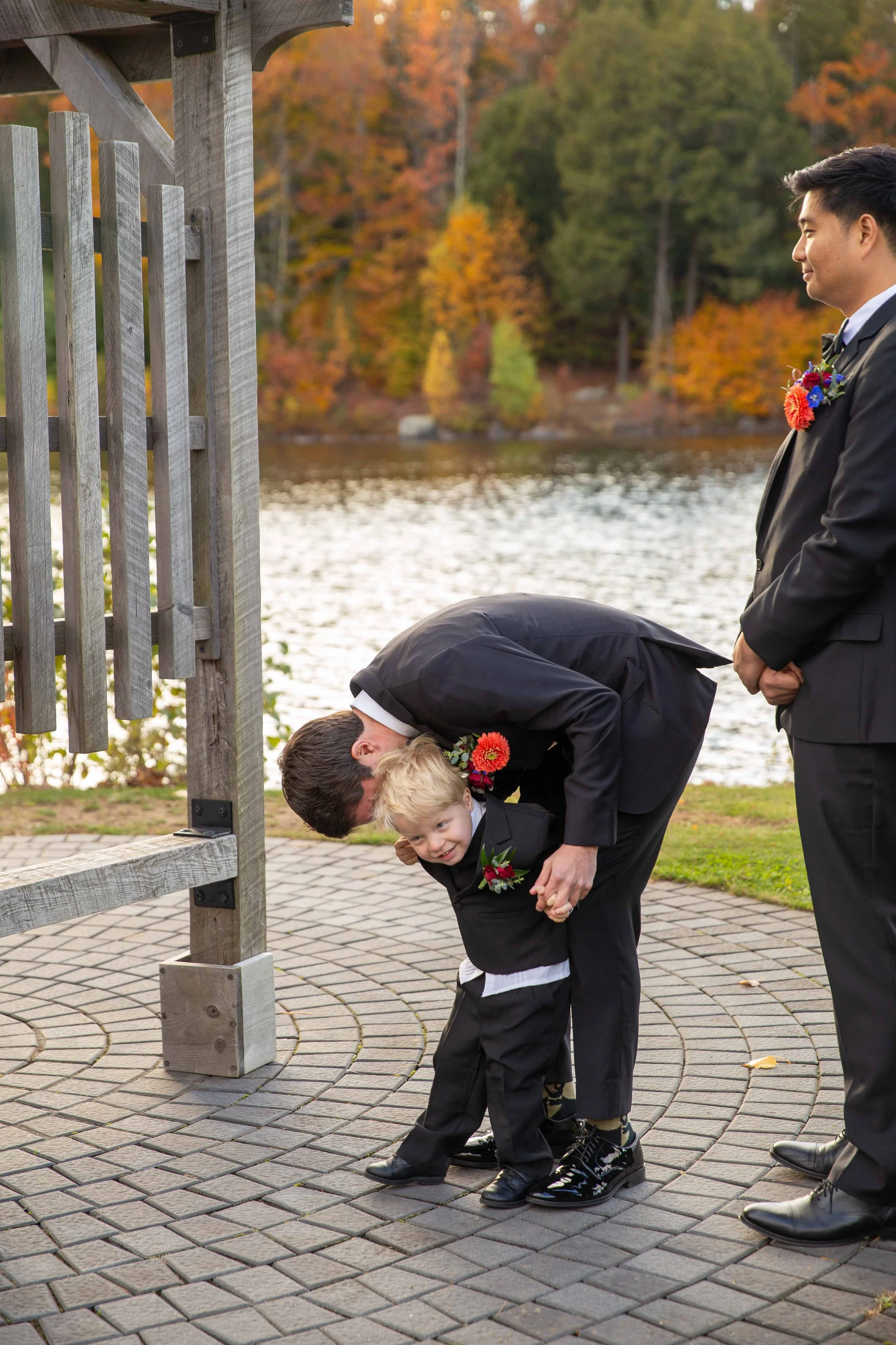 Two men in suits and a young boy in formal attire near a body of water with fall foliage in the background. One man is leaning down and kissing the boy on the head while the other man stands nearby with hands clasped.