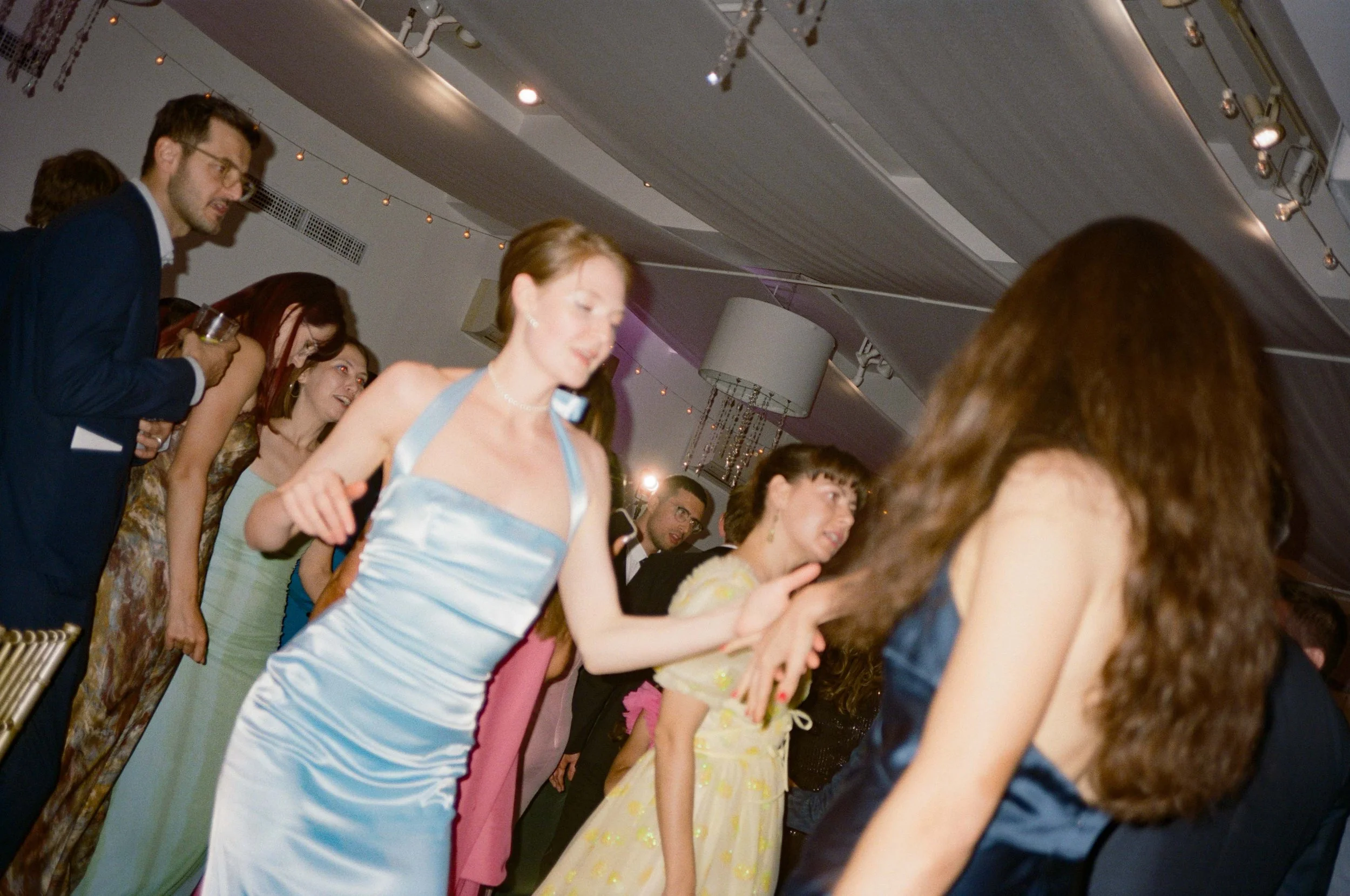 People dancing at a formal event, with women in elegant dresses and men in suits, in a decorated indoor venue.