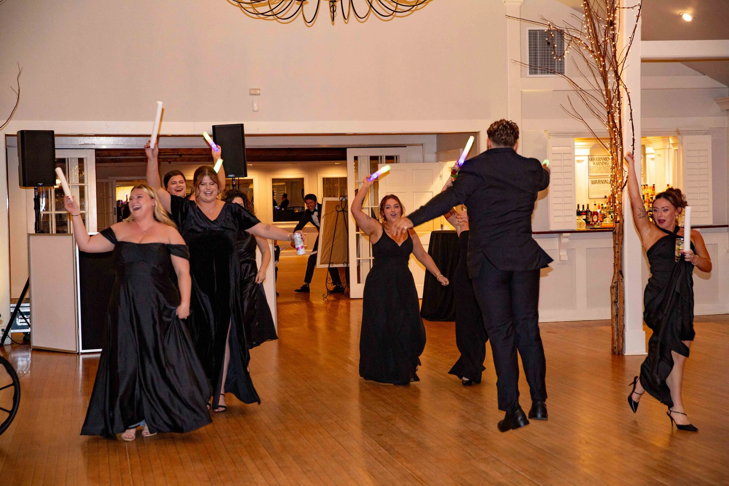 People dancing at a formal event, some holding glow sticks and drinks, in a decorated banquet hall.