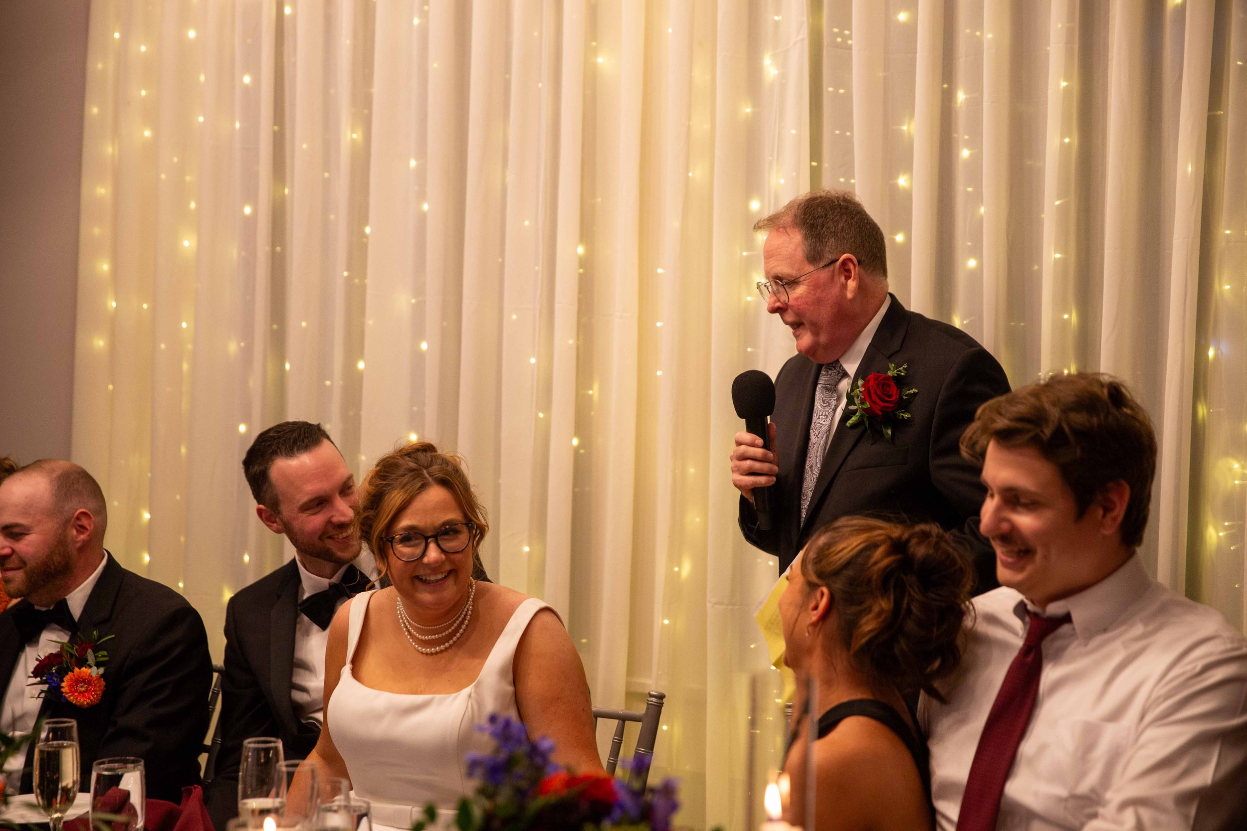 A man in a suit with a red rose boutonniere giving a speech at a wedding reception, with guests seated at a table and a white curtain with string lights in the background.