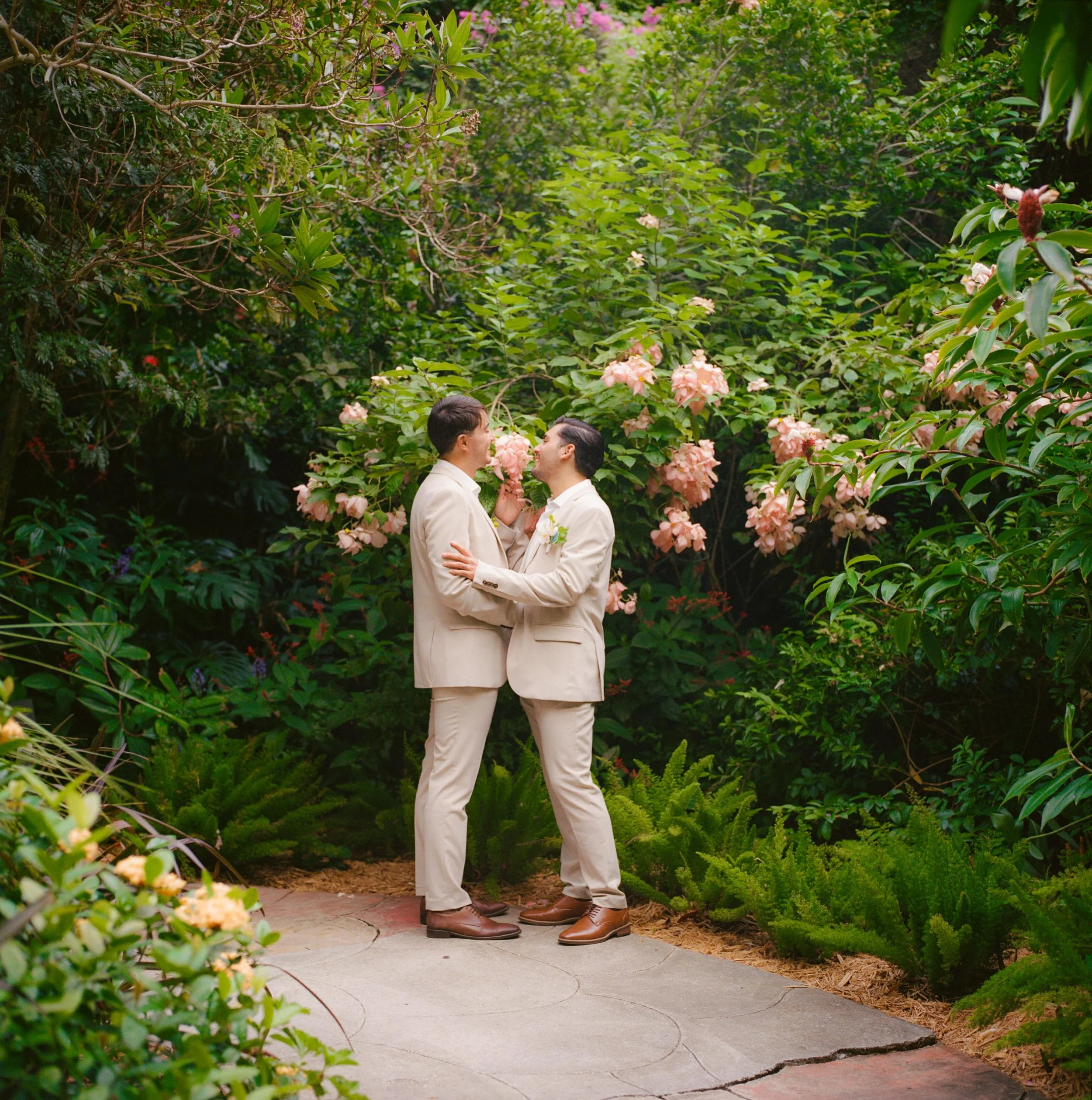 Two men in beige suits holding hands and smiling at each other in a lush garden with pink flowers and green foliage.