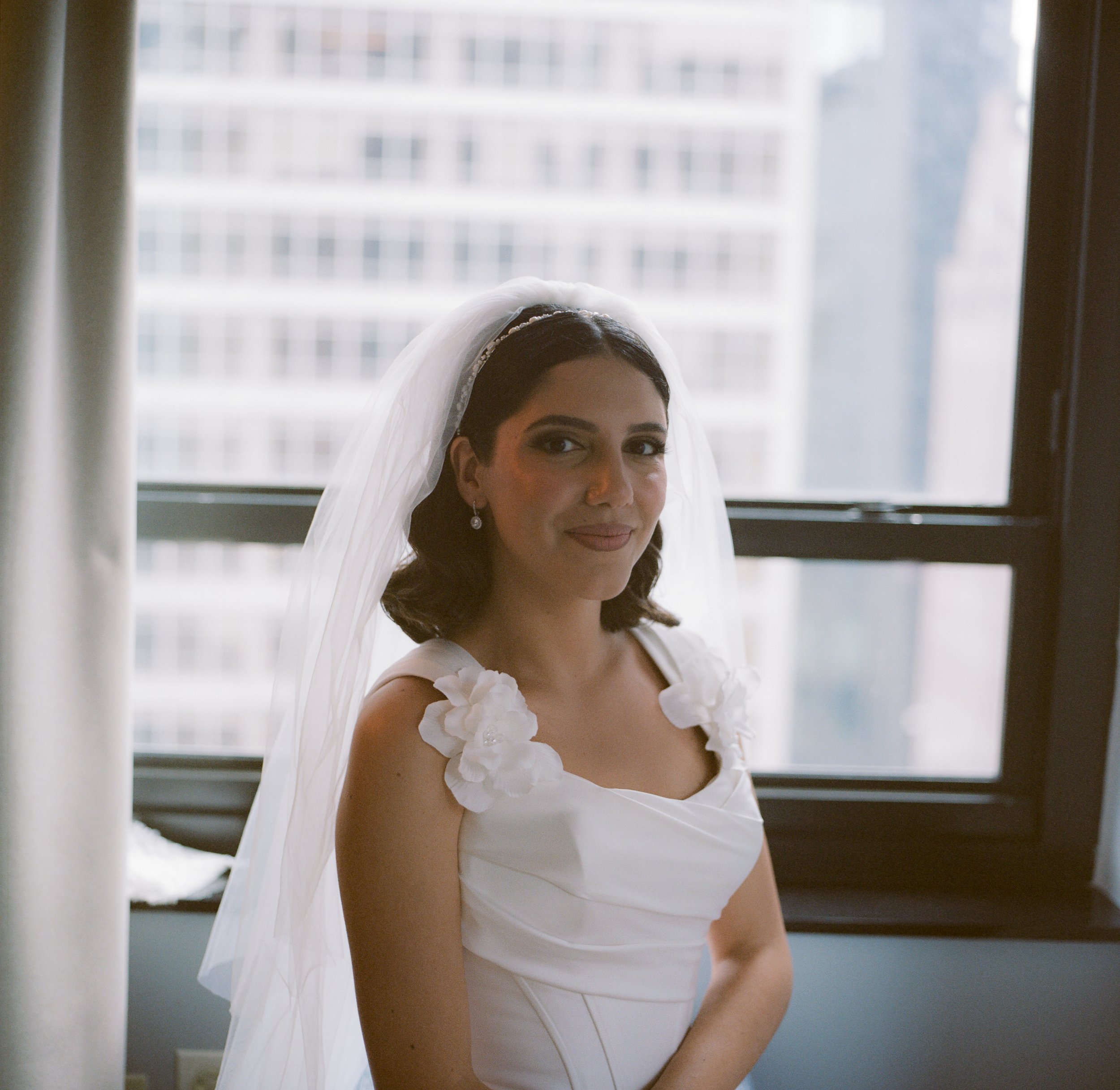 A bride with dark hair, wearing a white wedding dress with floral details on the shoulders, and a veil, standing in front of a window with urban buildings outside.