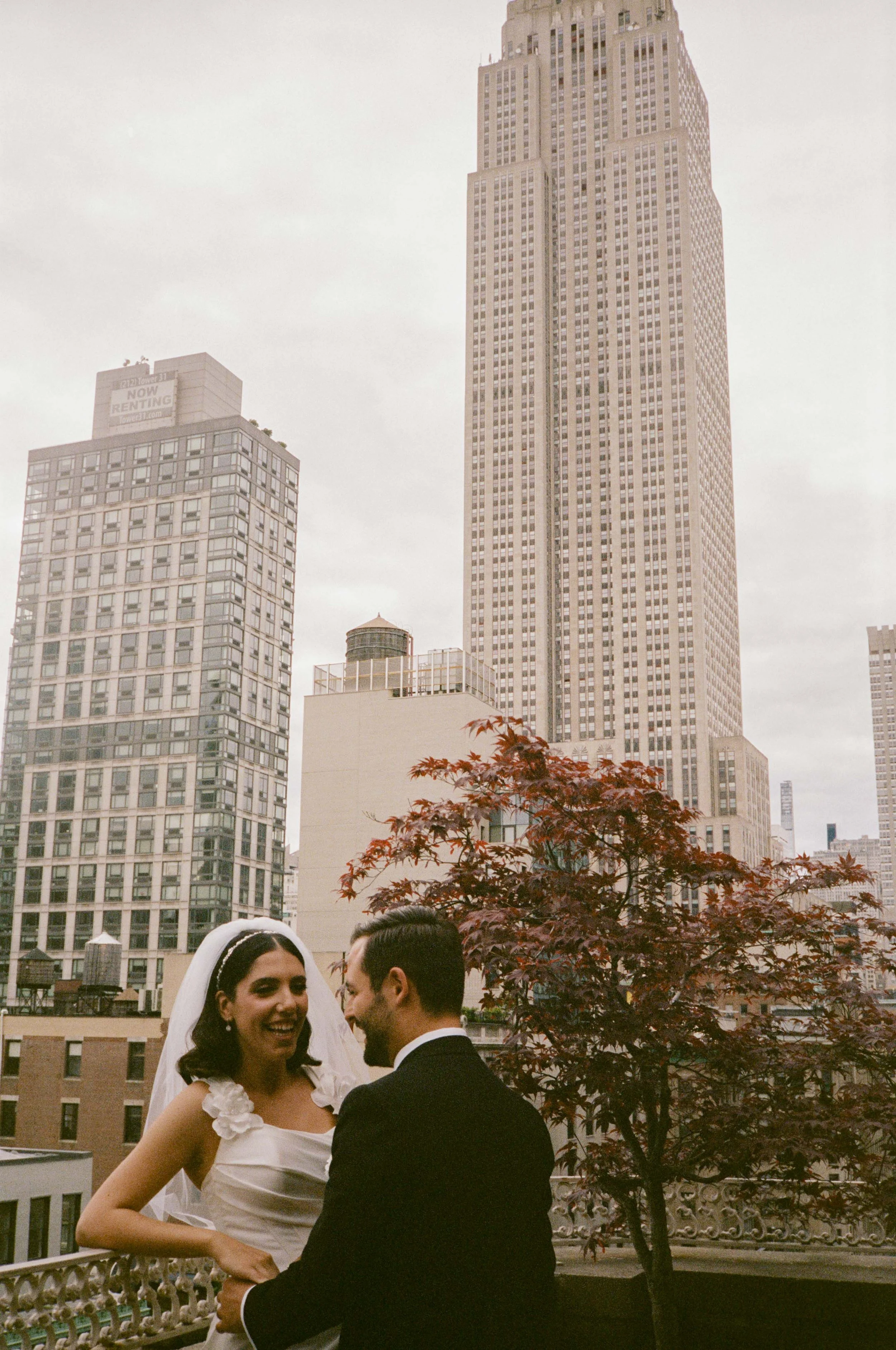 A bride and groom smiling on a balcony with New York City skyscrapers in the background.