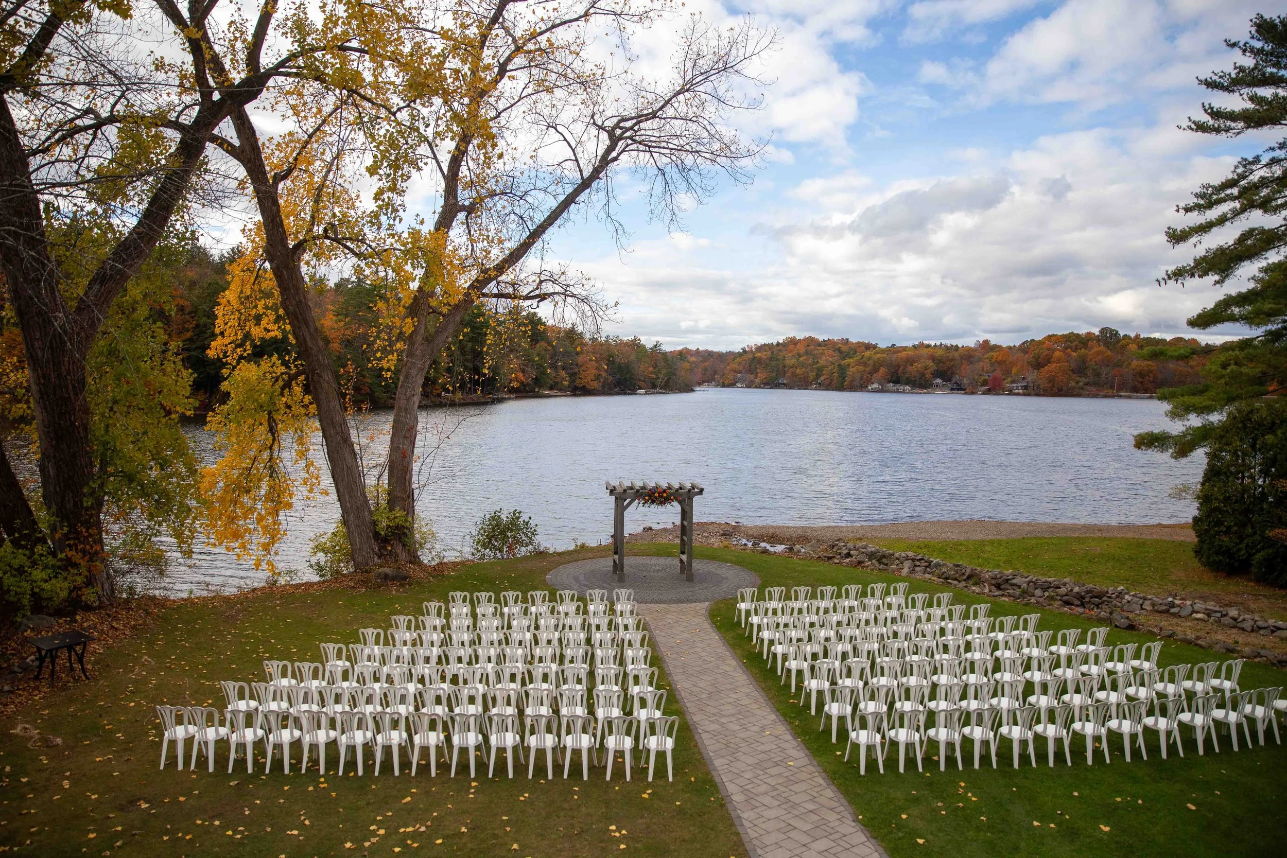 An outdoor wedding setup by a lake with rows of white chairs leading to an altar, surrounded by autumn trees and a partly cloudy sky.