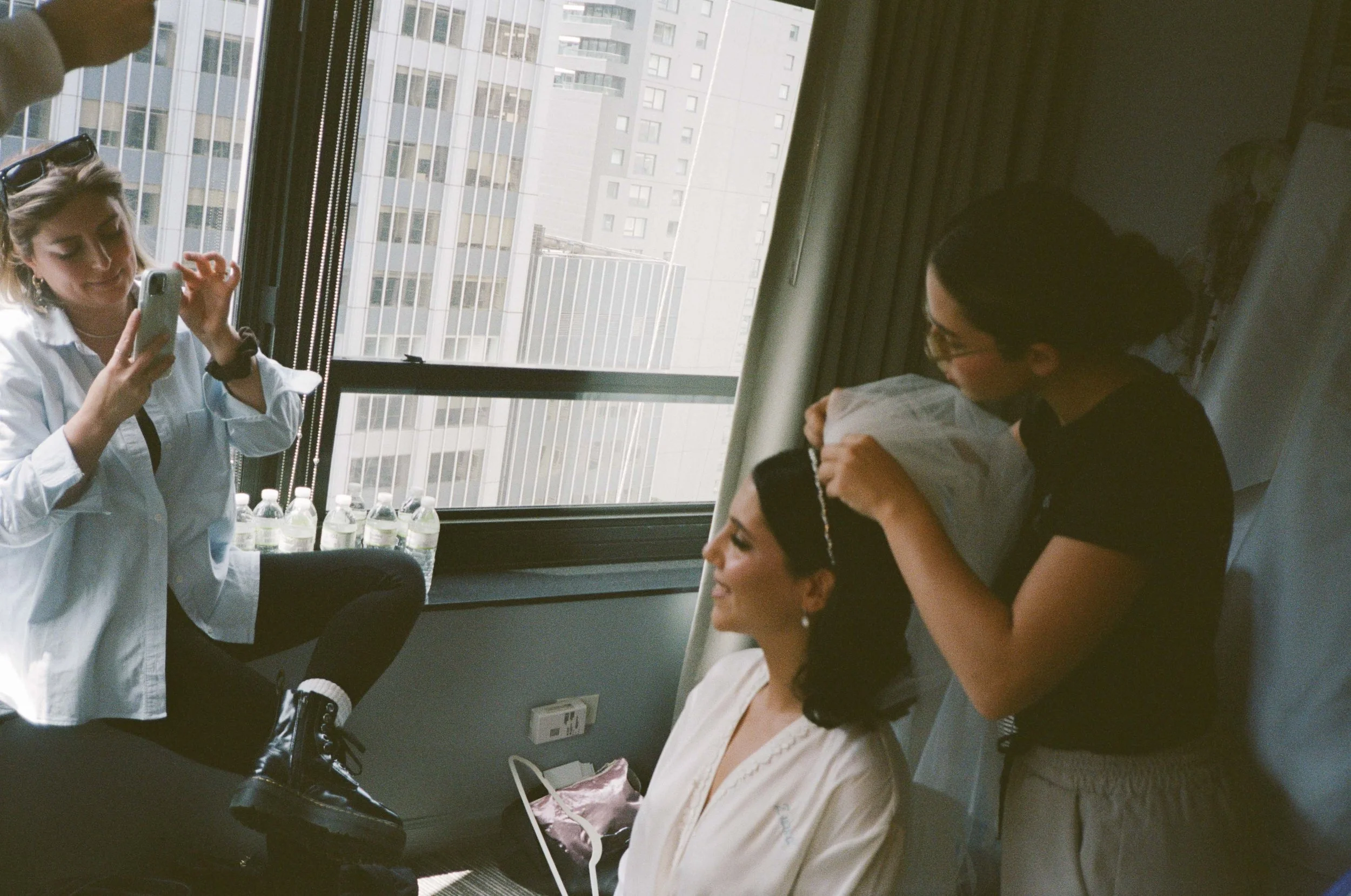 A woman sitting on a bed with her eyes closed, smiling as another woman styles her hair with a headband, while a third woman takes a photo with a smartphone in a hotel room with a large window showing city buildings.