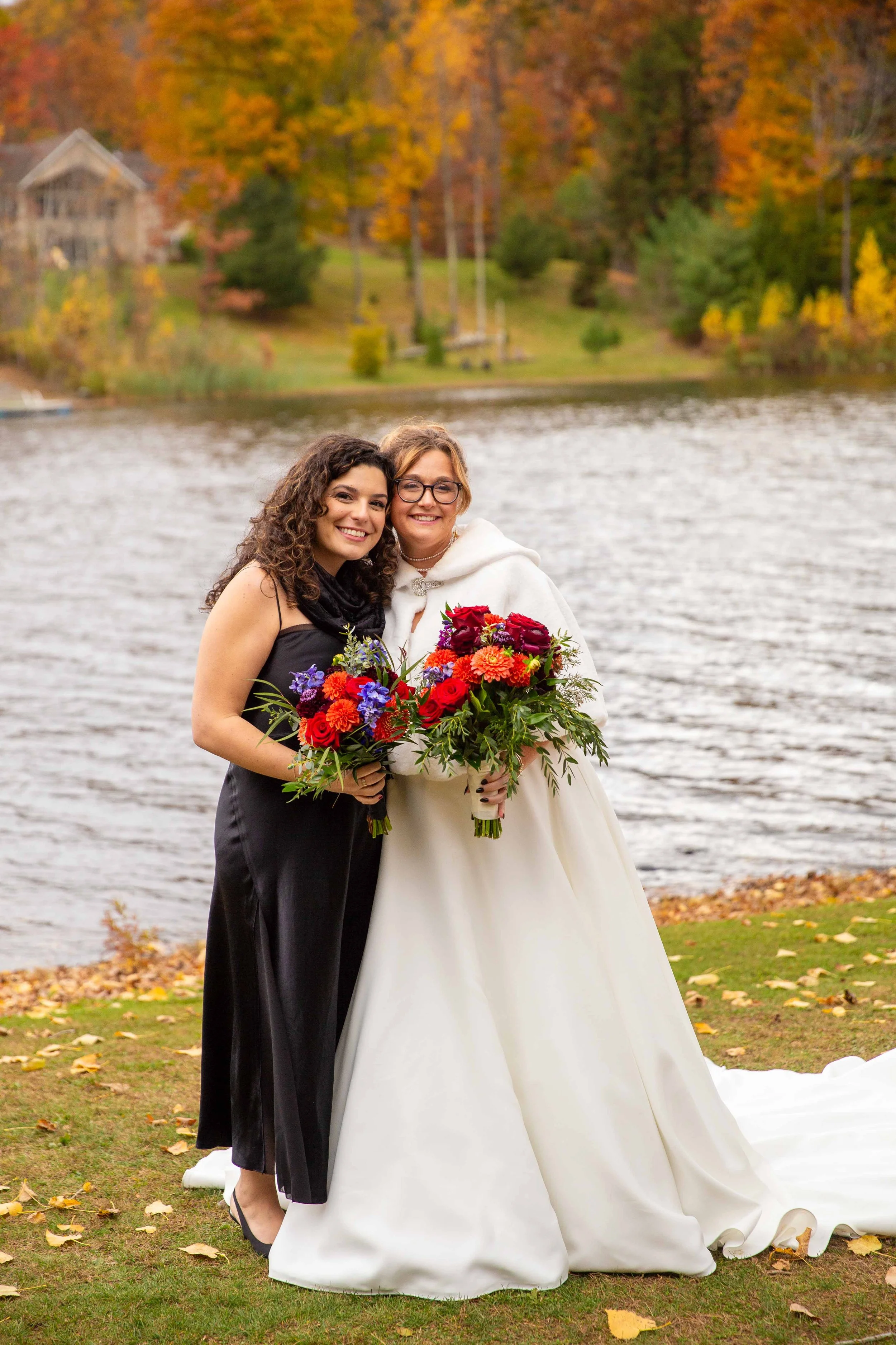 Two women, one in a white wedding dress and the other in a black dress, standing by a lake holding bouquets of colorful flowers with an autumn landscape in the background.