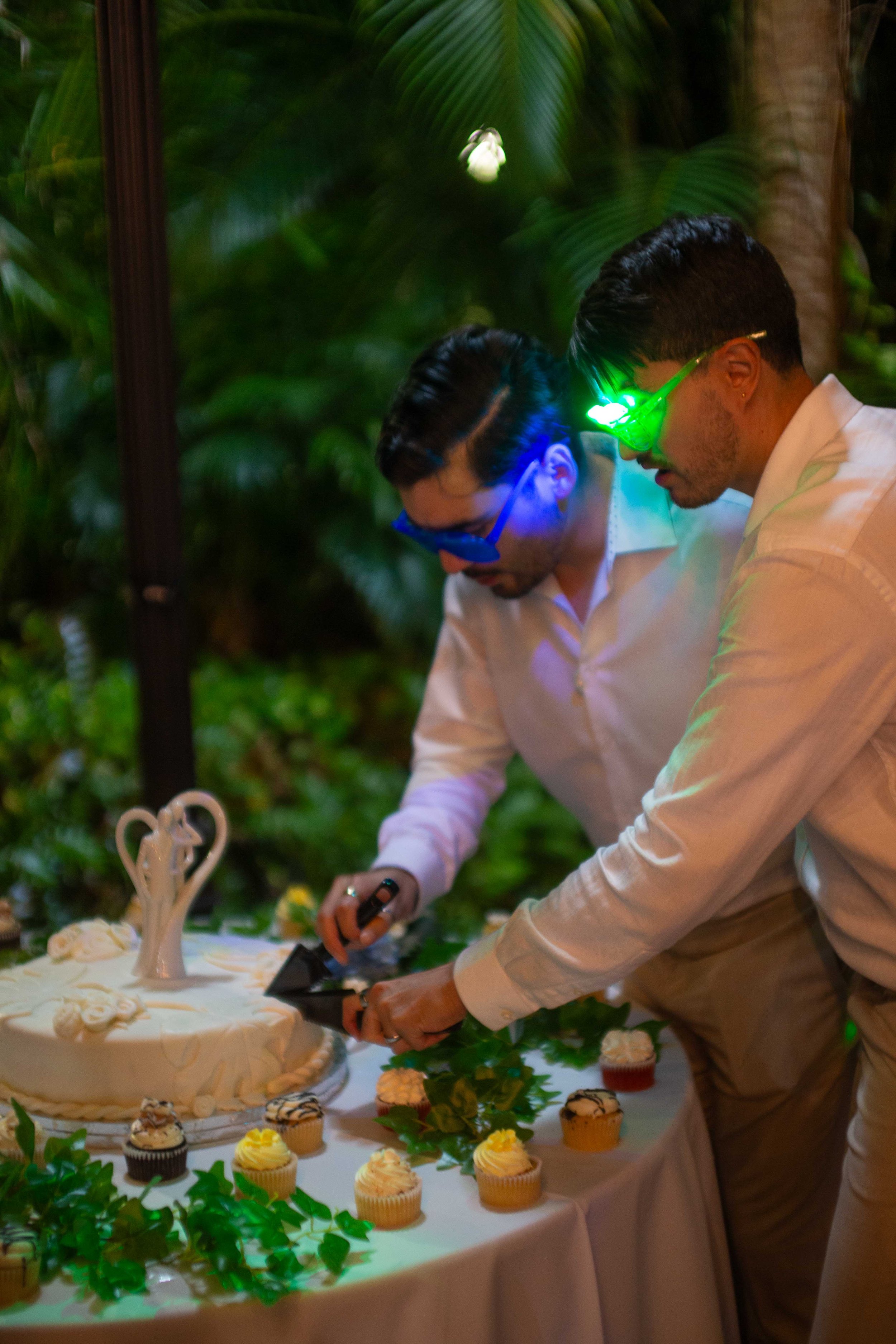 Two men with sunglasses cutting a wedding cake decorated with a swan topper, surrounded by cupcakes and green foliage, outdoors at night.