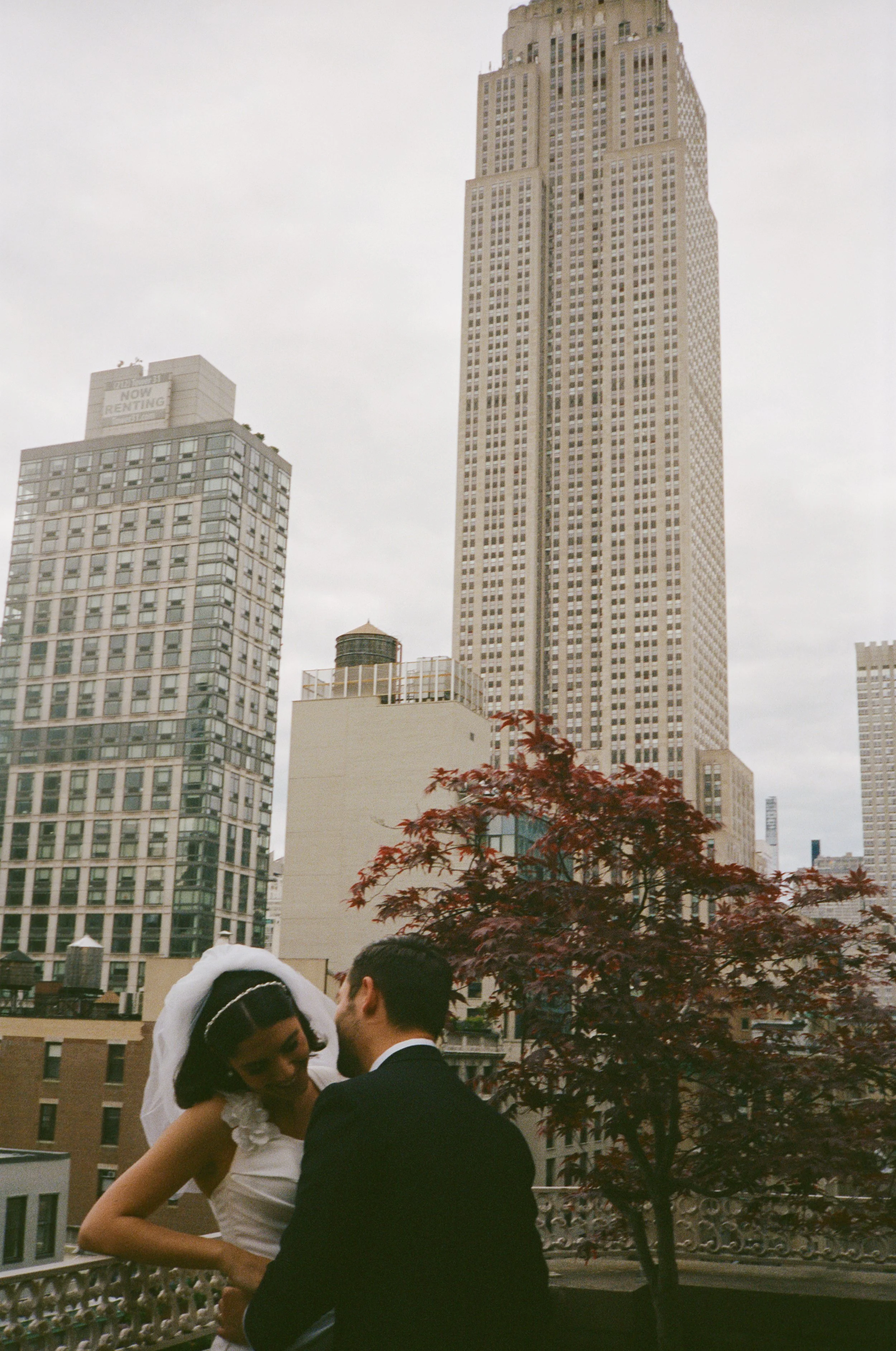 A bride and groom on a rooftop with New York City skyscrapers in the background, including a prominent Art Deco building and a tree with reddish leaves.