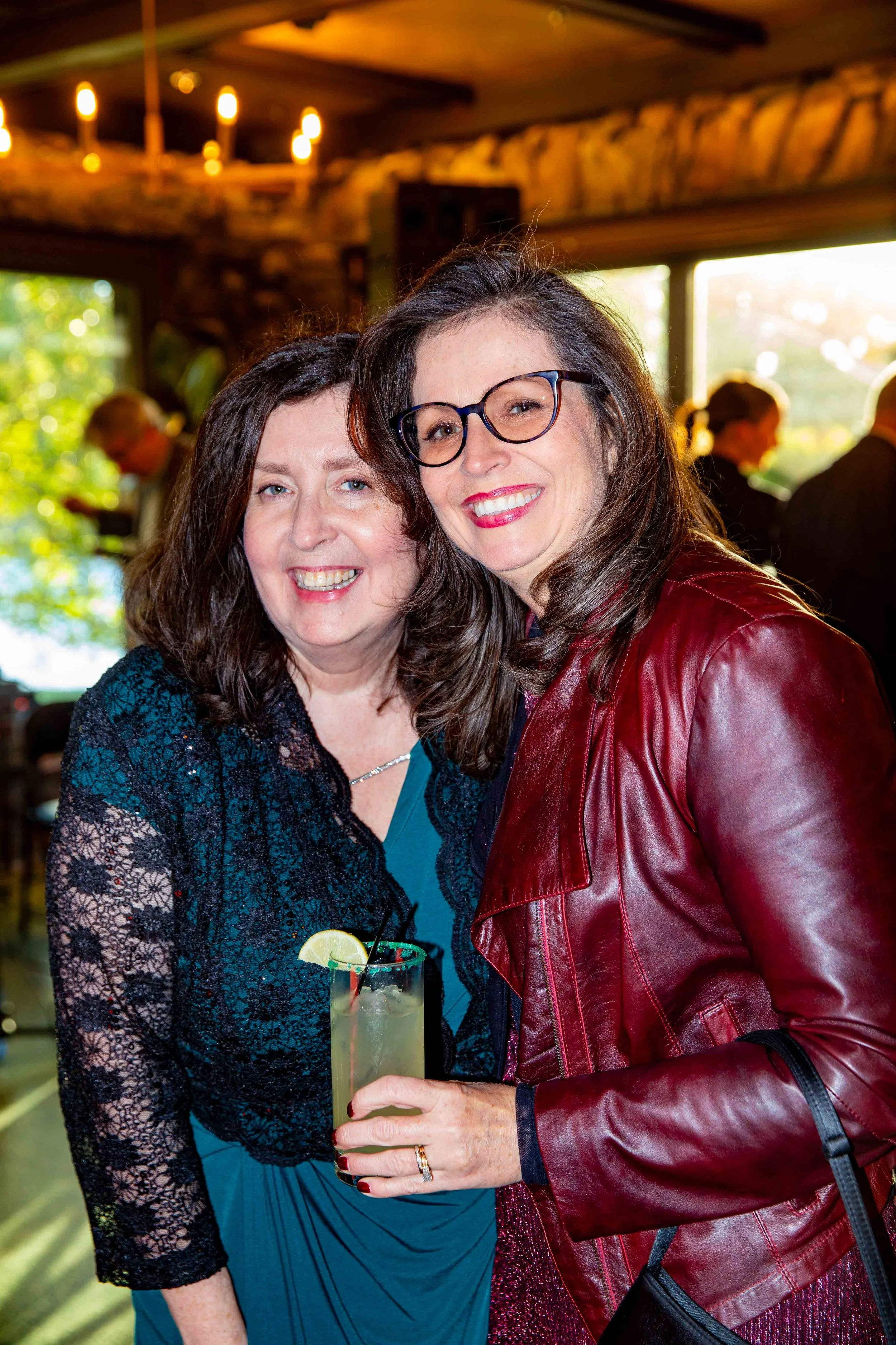 Two women smiling and enjoying a drink at a social gathering in a rustic indoor setting with large windows and warm lighting.