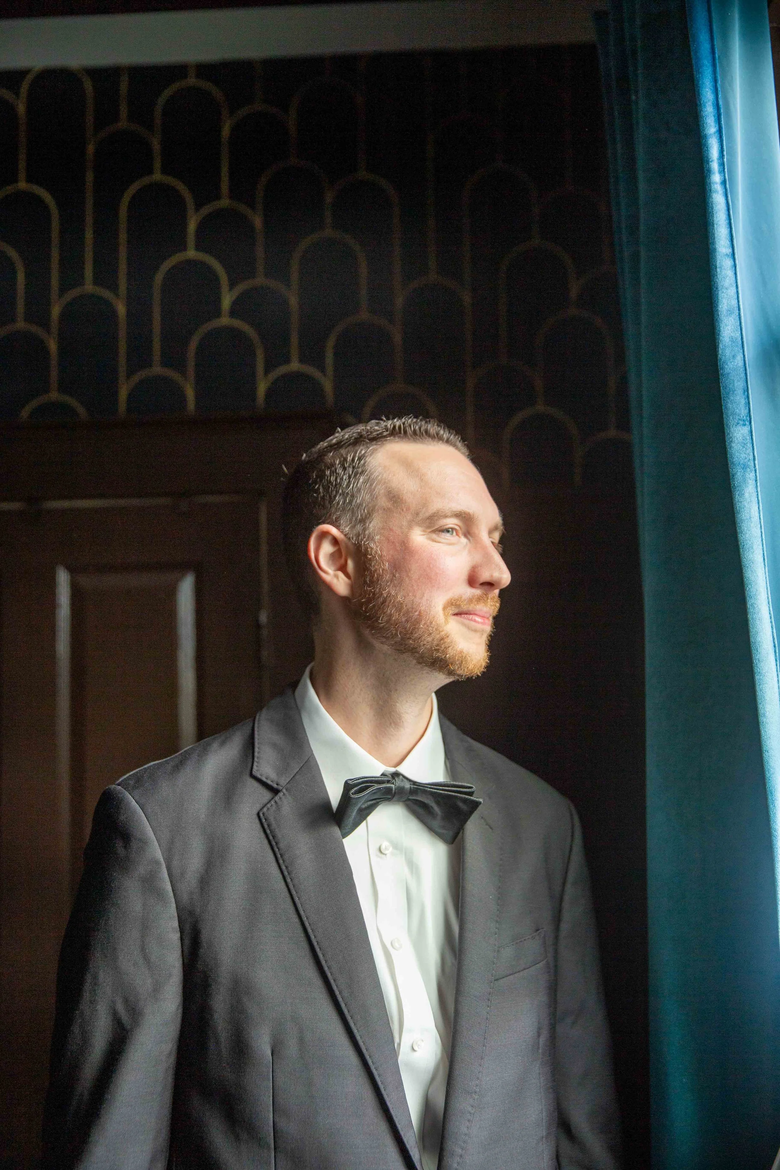A man dressed in a black tuxedo with a bow tie, looking out a window with sunlight on his face.