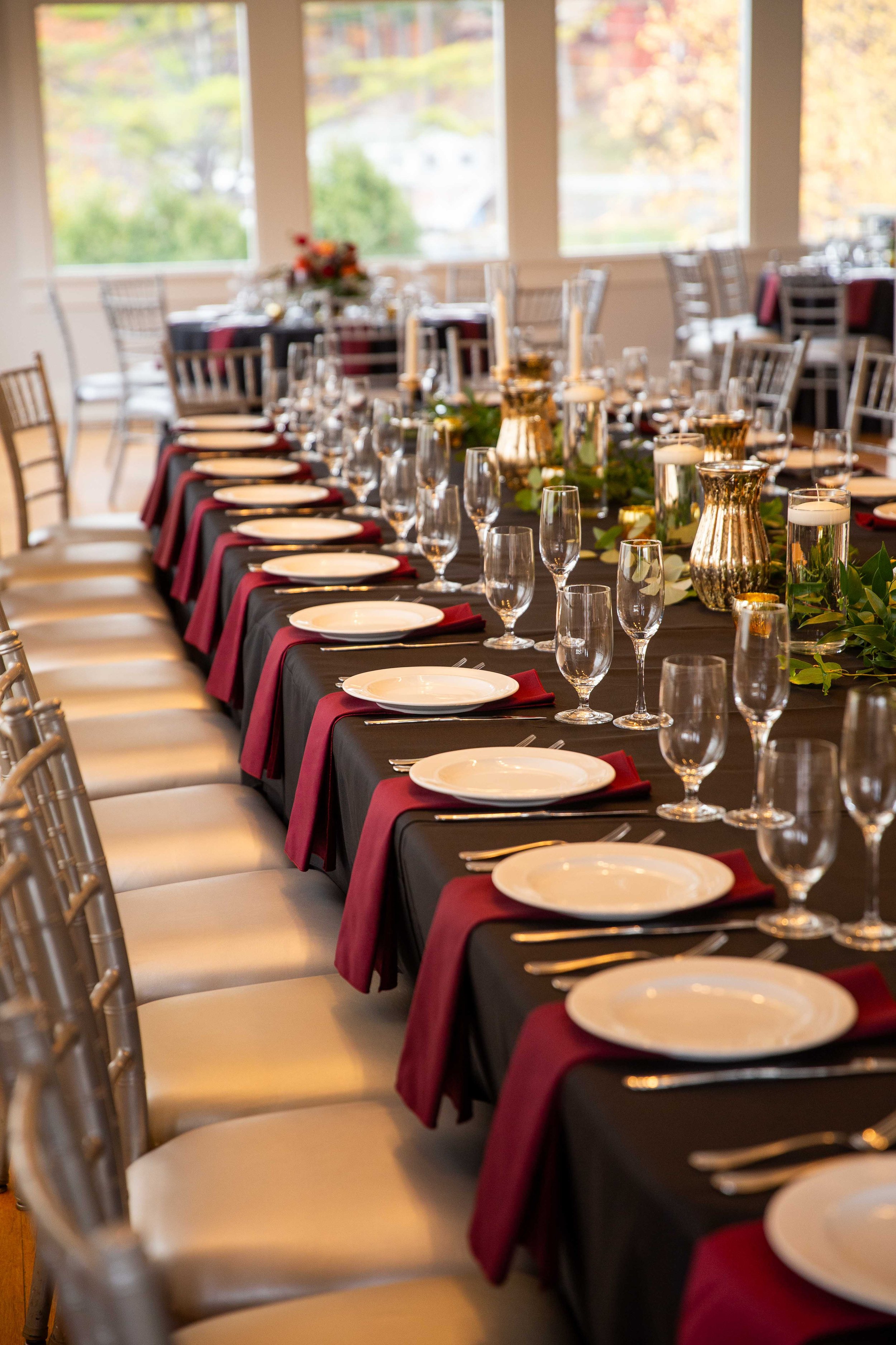Long dining table set for a formal event with white plates, silverware, red napkins, and empty glasses, decorated with candles and greenery, in a room with large windows and a view of trees with fall foliage.