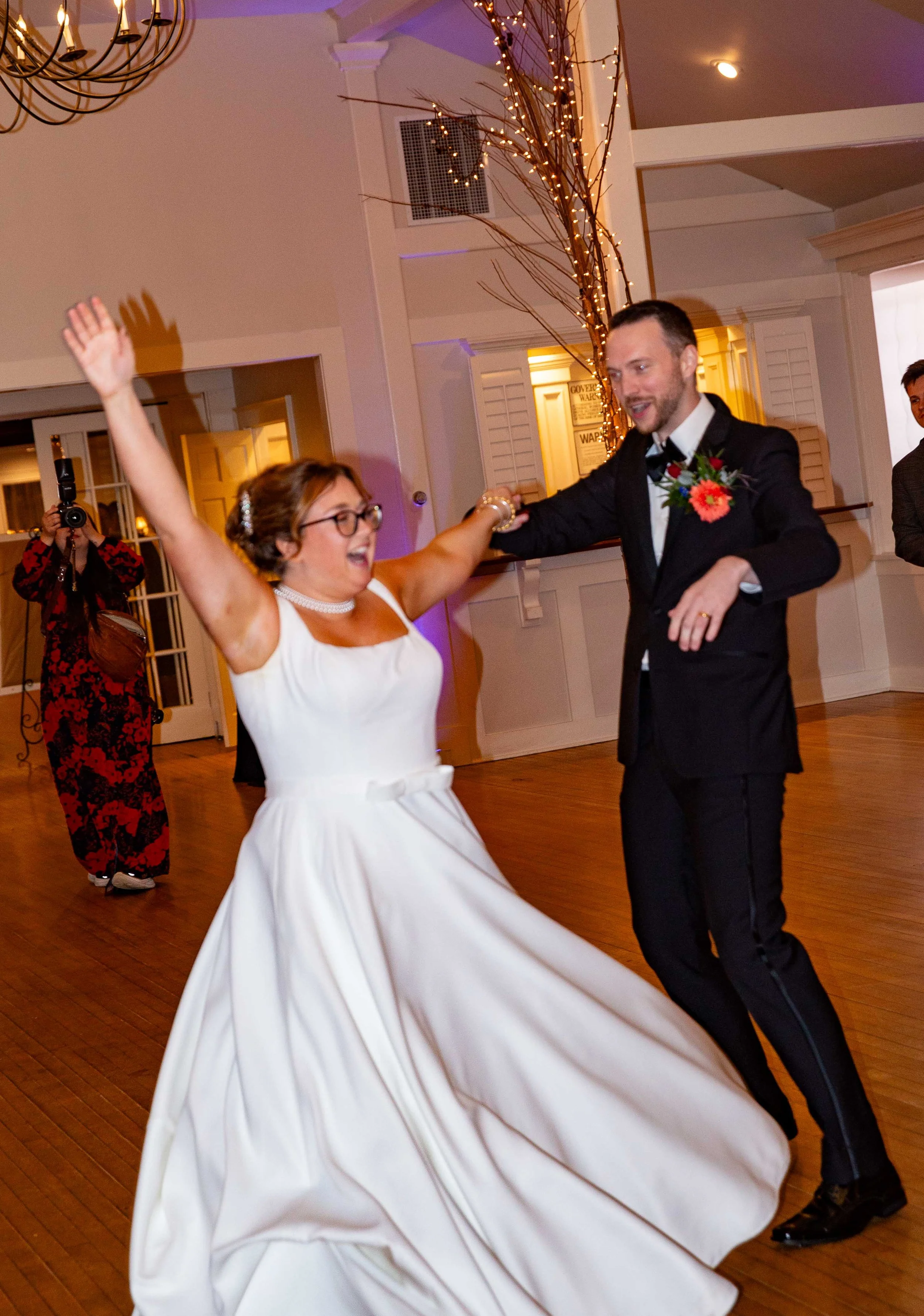 A bride and groom dancing happily at their wedding reception. The bride is wearing a white wedding dress and glasses, and the groom is in a black suit with a floral boutonniere. They are smiling and holding hands as they dance.