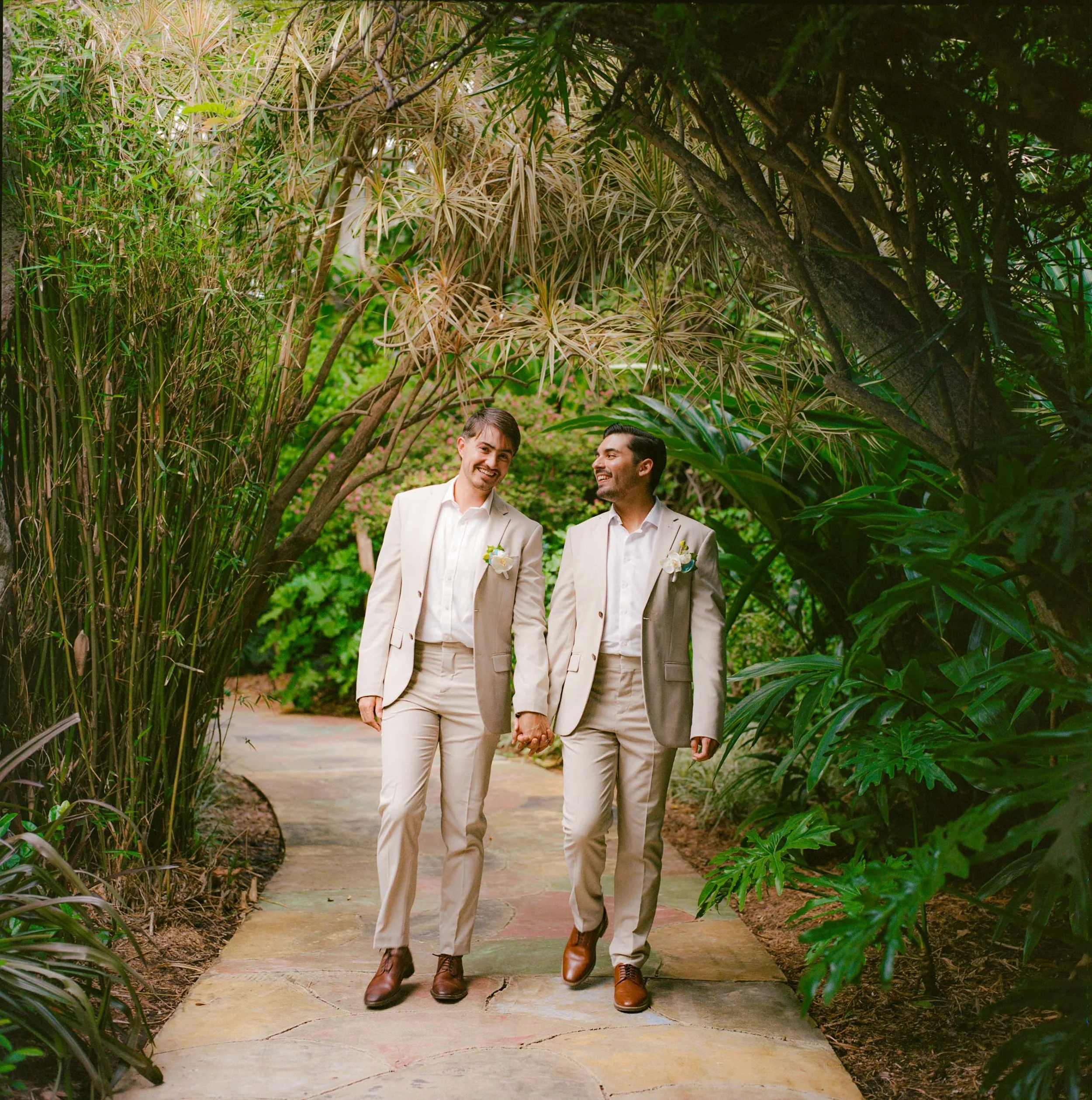Two men in beige suits holding hands and walking through a lush, green garden pathway, smiling at each other.