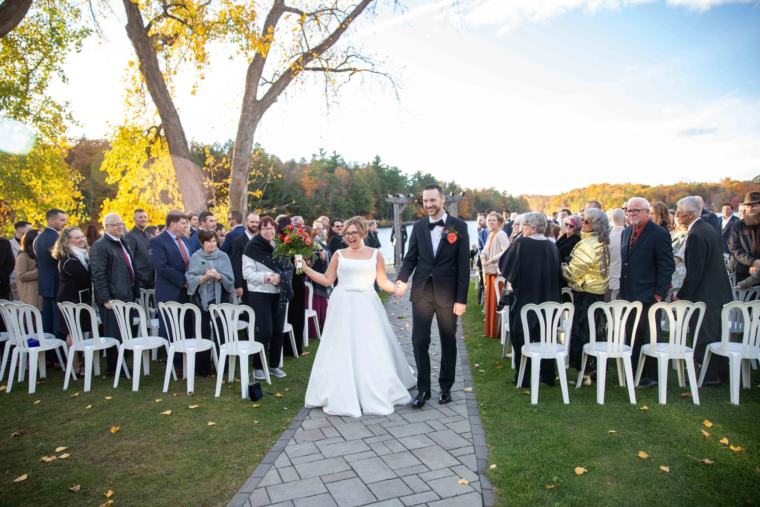 A newly married couple walking down the aisle outdoors during a wedding ceremony in the fall, with guests on either side of the path.