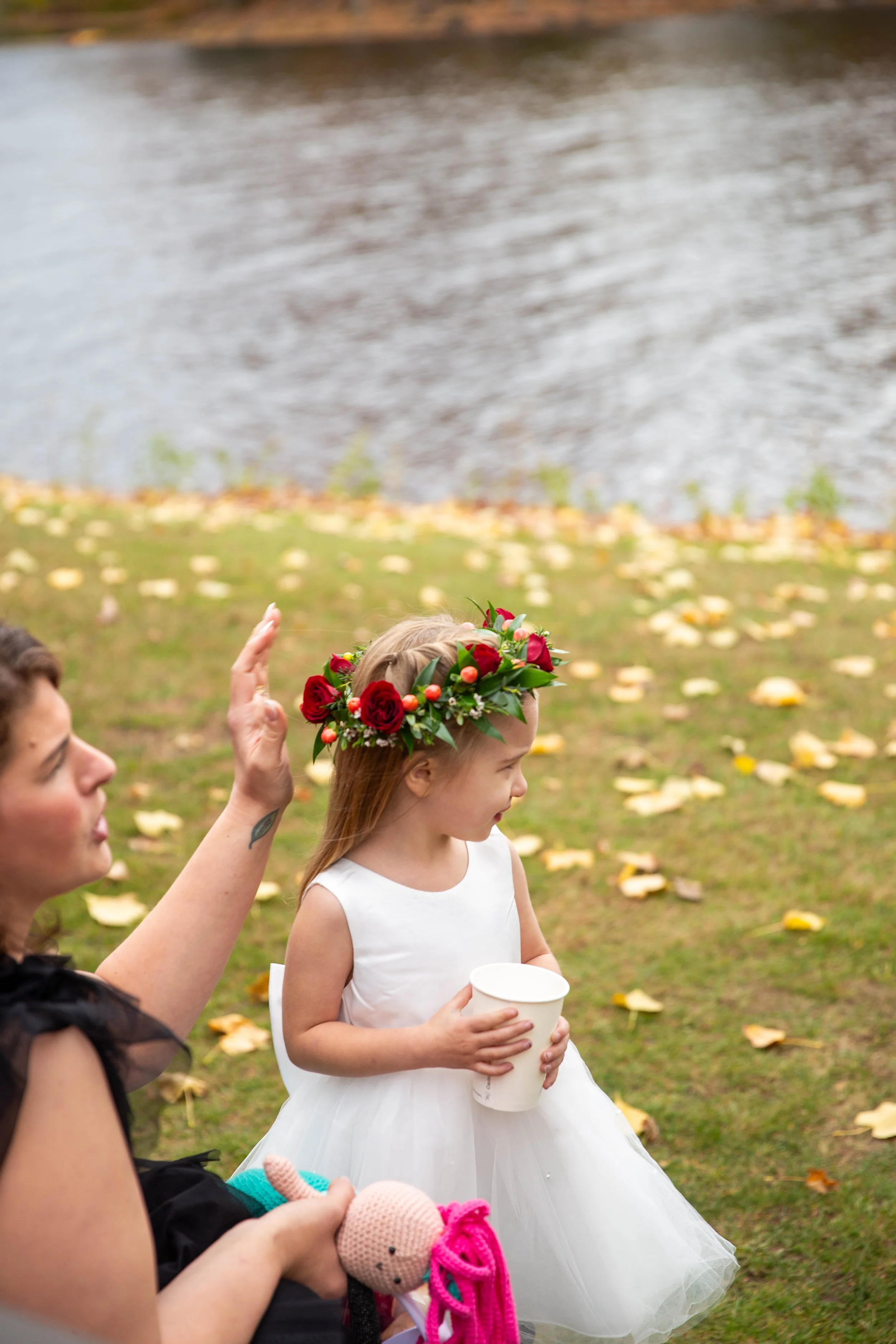 A young girl in a white dress with a floral crown made of red roses is holding a paper cup, standing outdoors near a body of water surrounded by fallen autumn leaves. An adult woman beside her is waving and holding a stuffed toy.