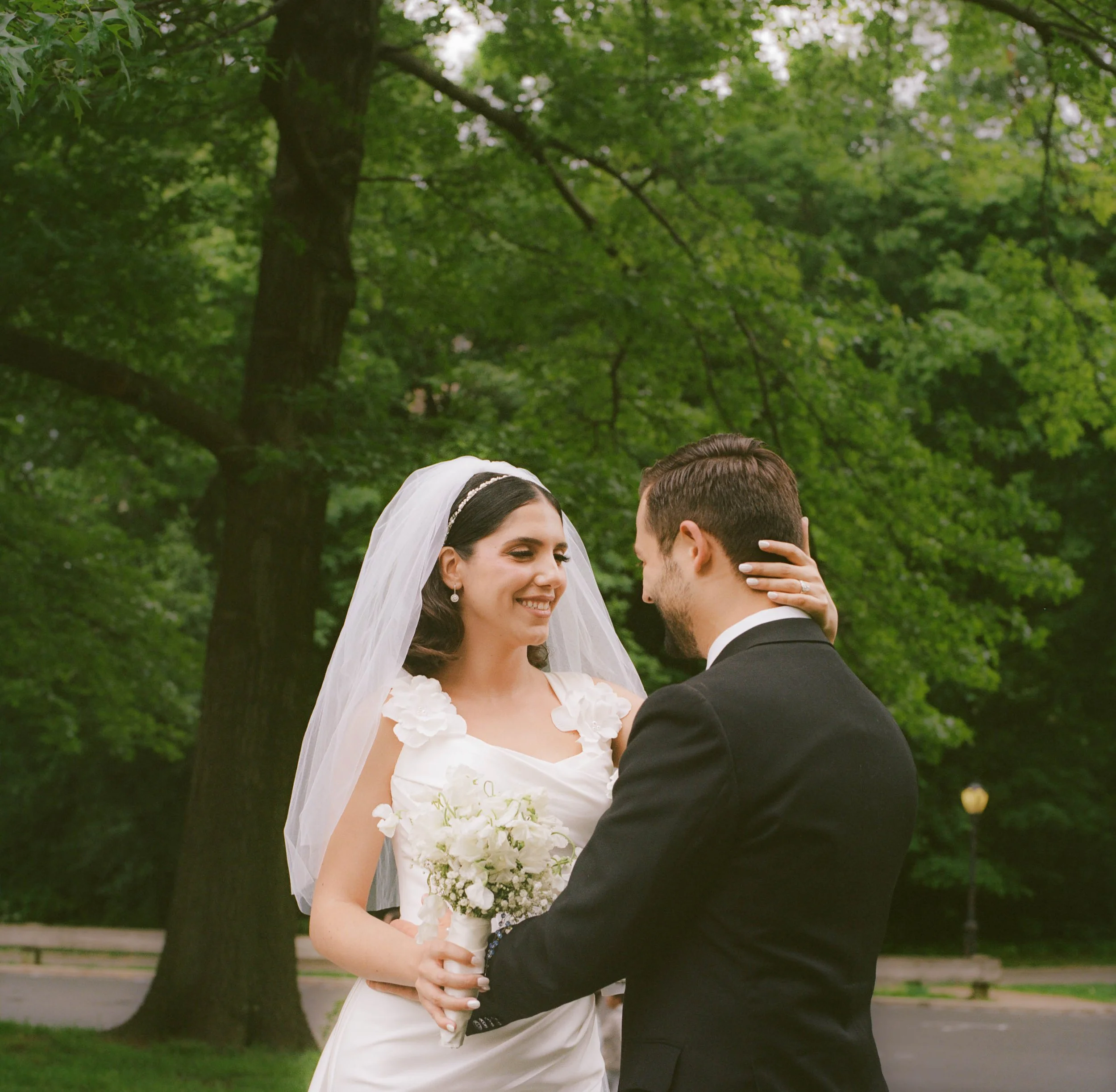 A bride and groom smiling and holding a bouquet of white flowers, standing outdoors under green trees.