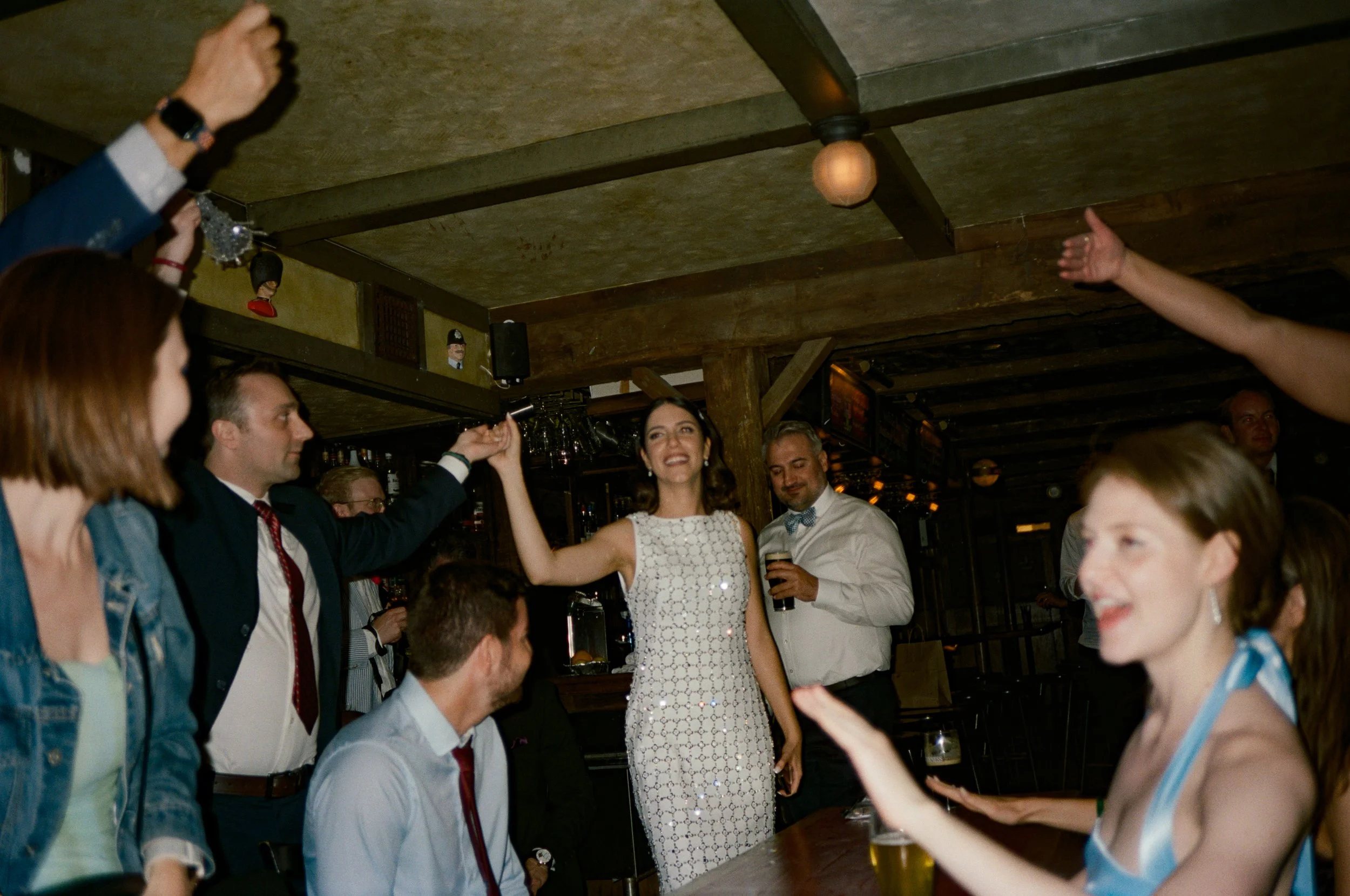 People celebrating at a party, with a woman in a white dress dancing and smiling, others clapping and holding drinks, in a wooden decorated venue.