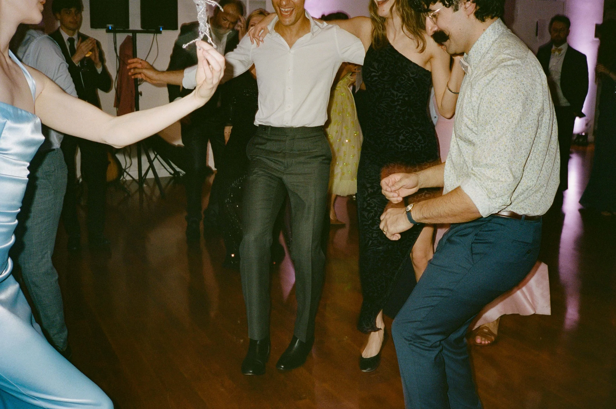 People dancing and having fun at a party or celebration indoors. The image shows a woman in a blue dress, a man in a light shirt, and others in the background enjoying the music and dance.