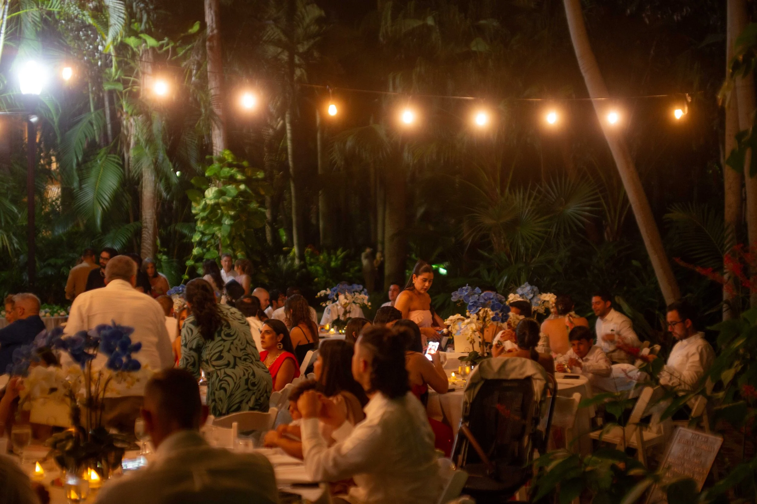 Guests dining at outdoor event under string lights surrounded by greenery and tropical plants at night.