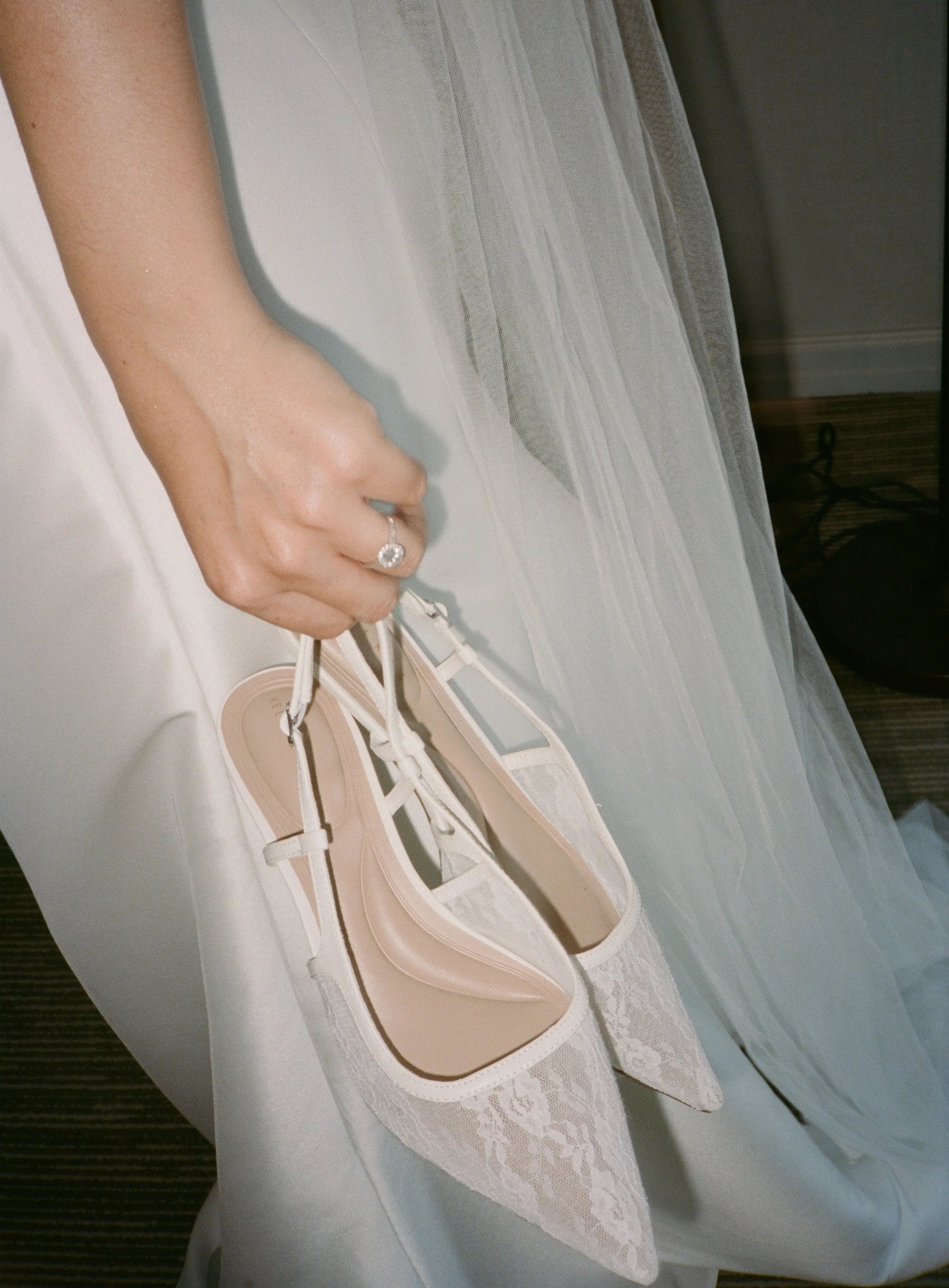 A bride holding a pair of white lace high heels, with a wedding dress in the background.