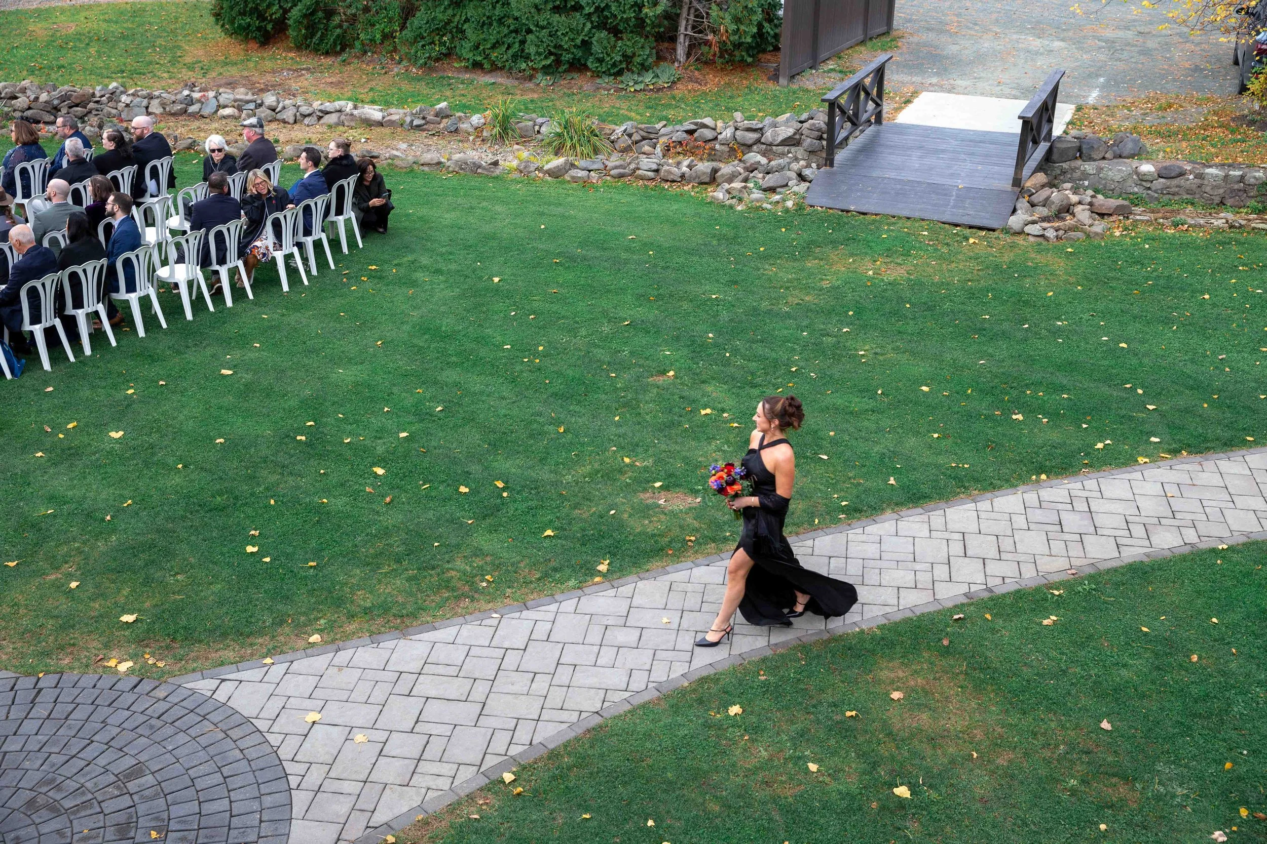 A woman in a black dress walking on a paved pathway holding a bouquet of flowers at an outdoor gathering, with guests seated at tables in the background.