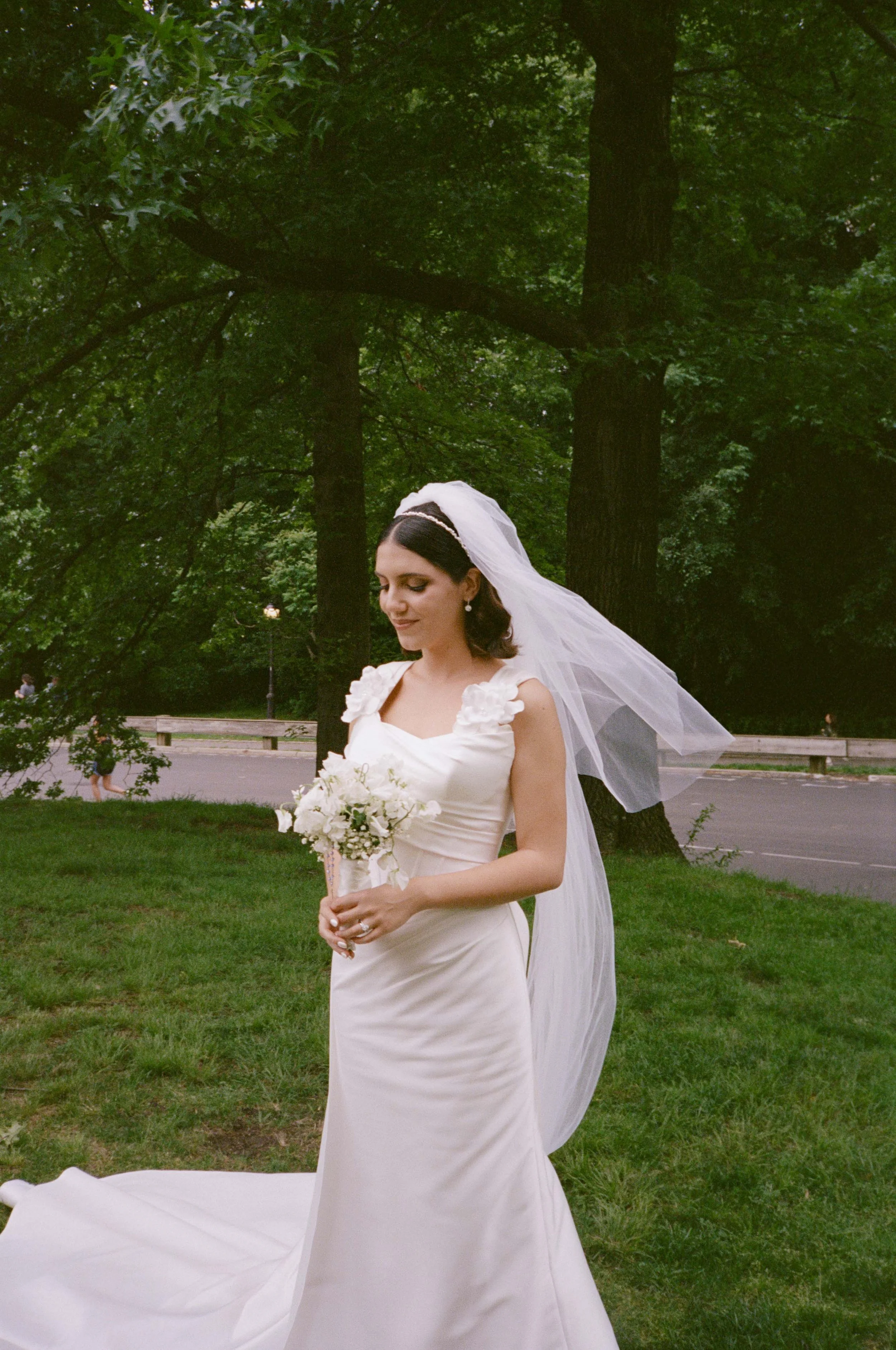 A bride in a white wedding gown holding a bouquet of flowers outdoors near trees and a parkway.