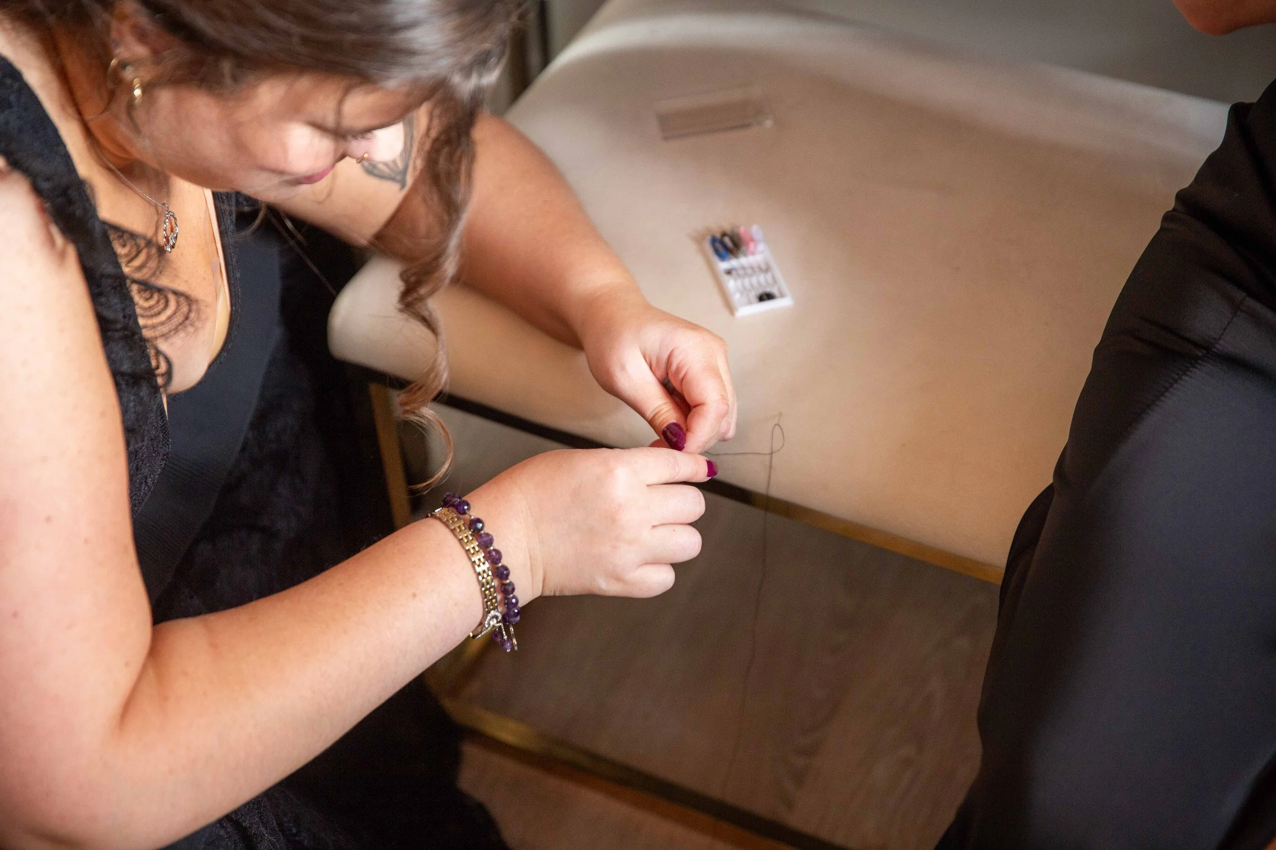 A woman with long hair working on sewing or embroidery, focusing on her hands with her needles and thread, sitting at a wooden desk.