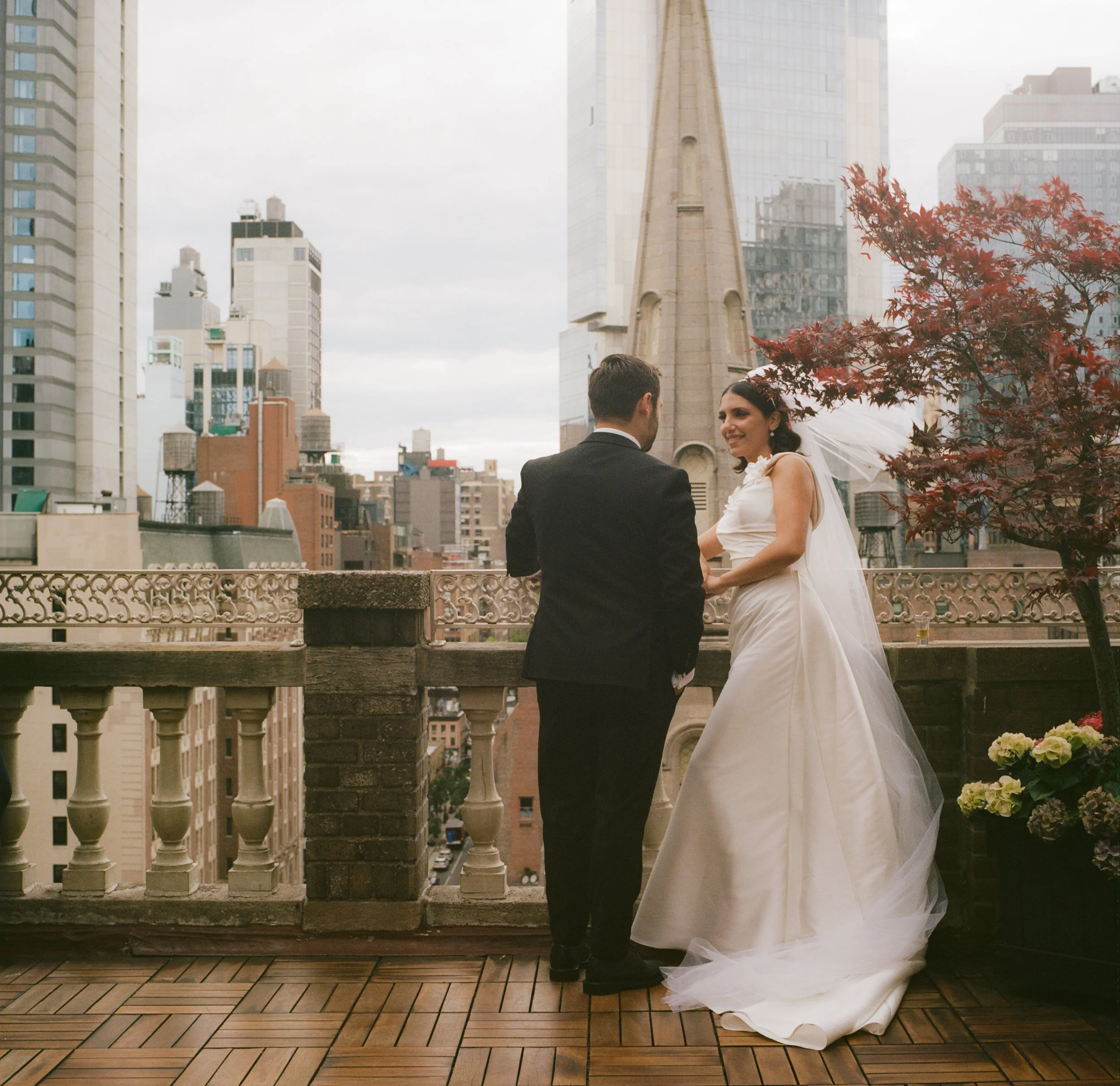 A bride and groom holding hands on a rooftop balcony with a city skyline in the background.