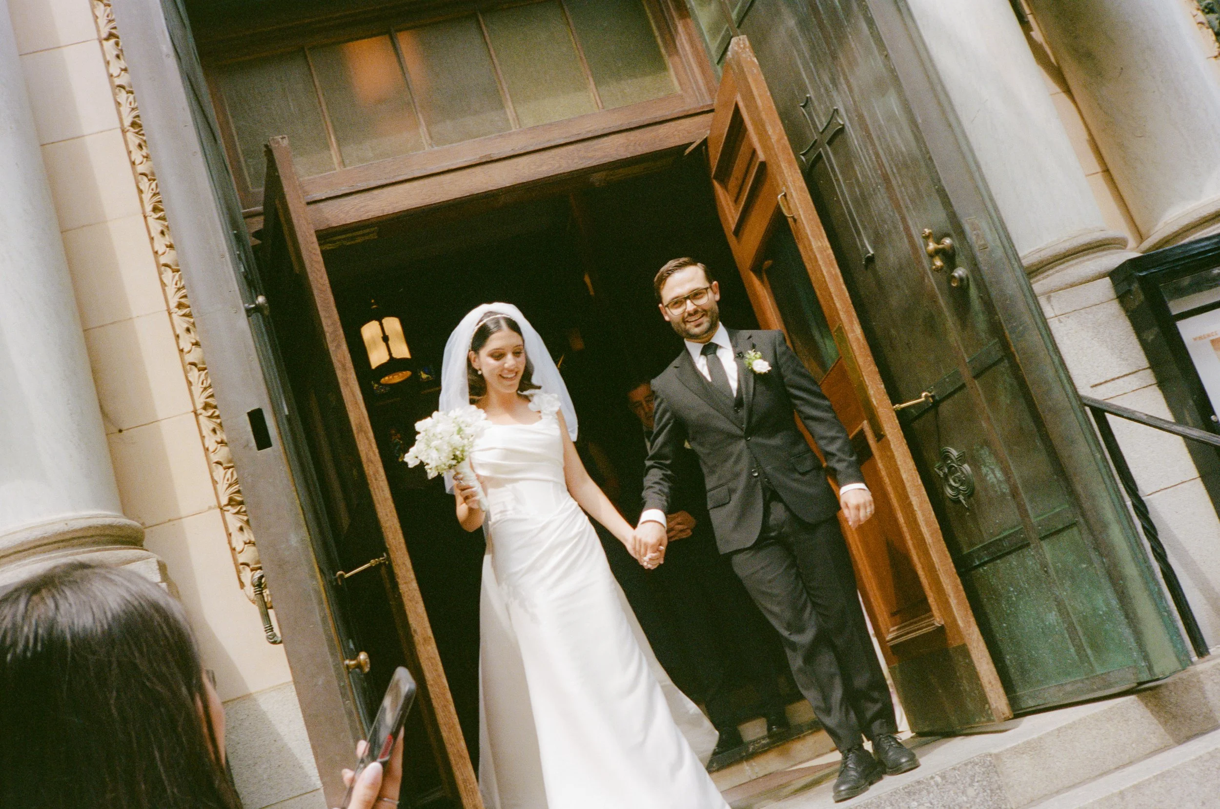 A newlywed couple exiting a church, holding hands and smiling. The bride is dressed in a white wedding gown and veil, holding a bouquet. The groom is in a black suit, white shirt, and tie, with glasses and a boutonniere. A person in the foreground is