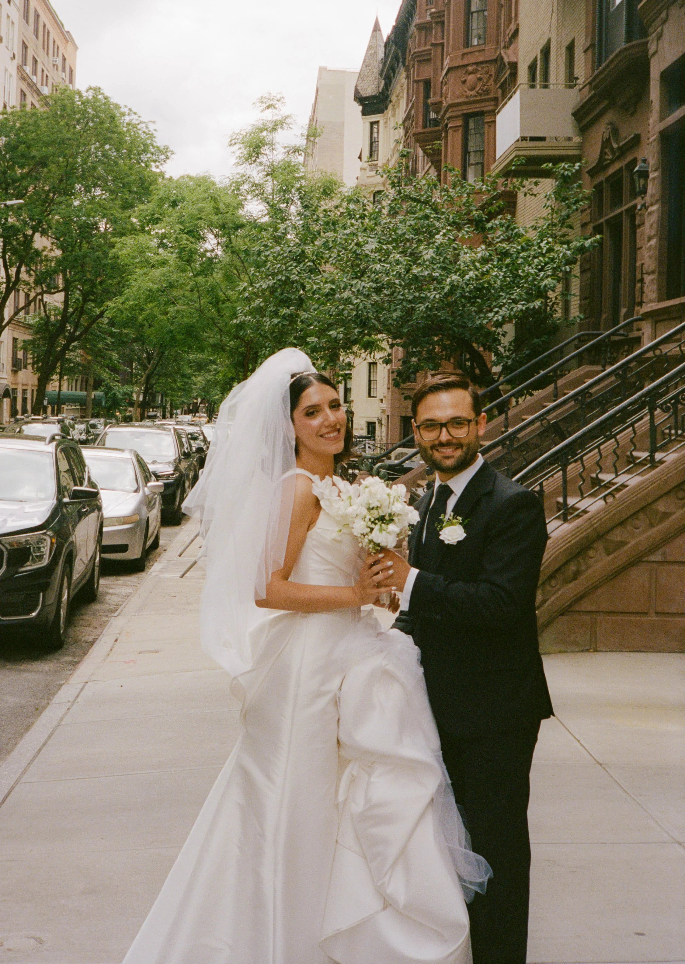 A bride and groom posing on a city sidewalk, with trees and brownstone buildings in the background. The bride is wearing a white wedding gown and veil, holding a bouquet of white flowers. The groom is in a black suit with a white shirt and black tie,