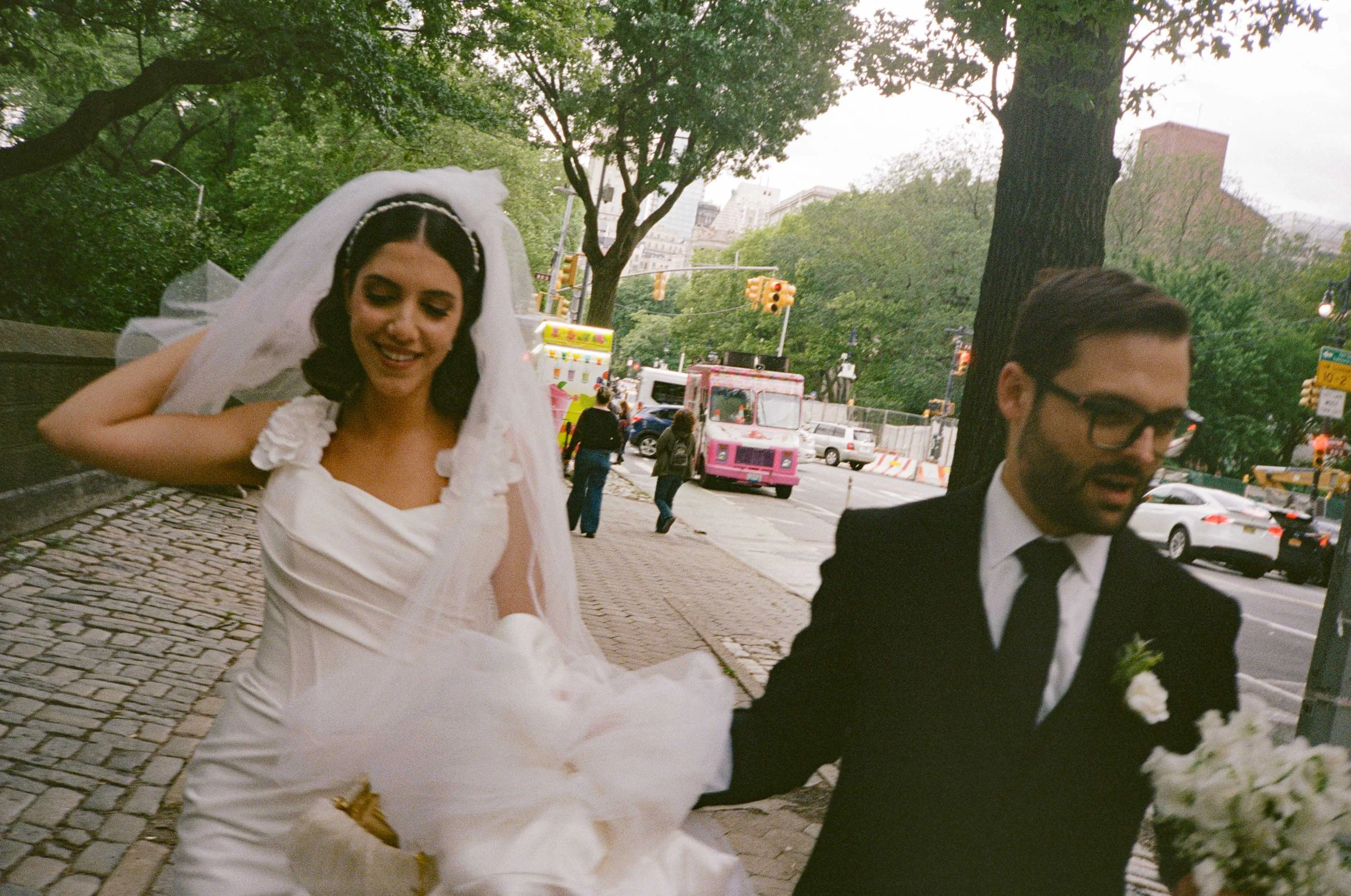 A bride in a white wedding dress and veil, smiling and holding her dress, walking with a groom in a black suit and glasses, on a city sidewalk.
