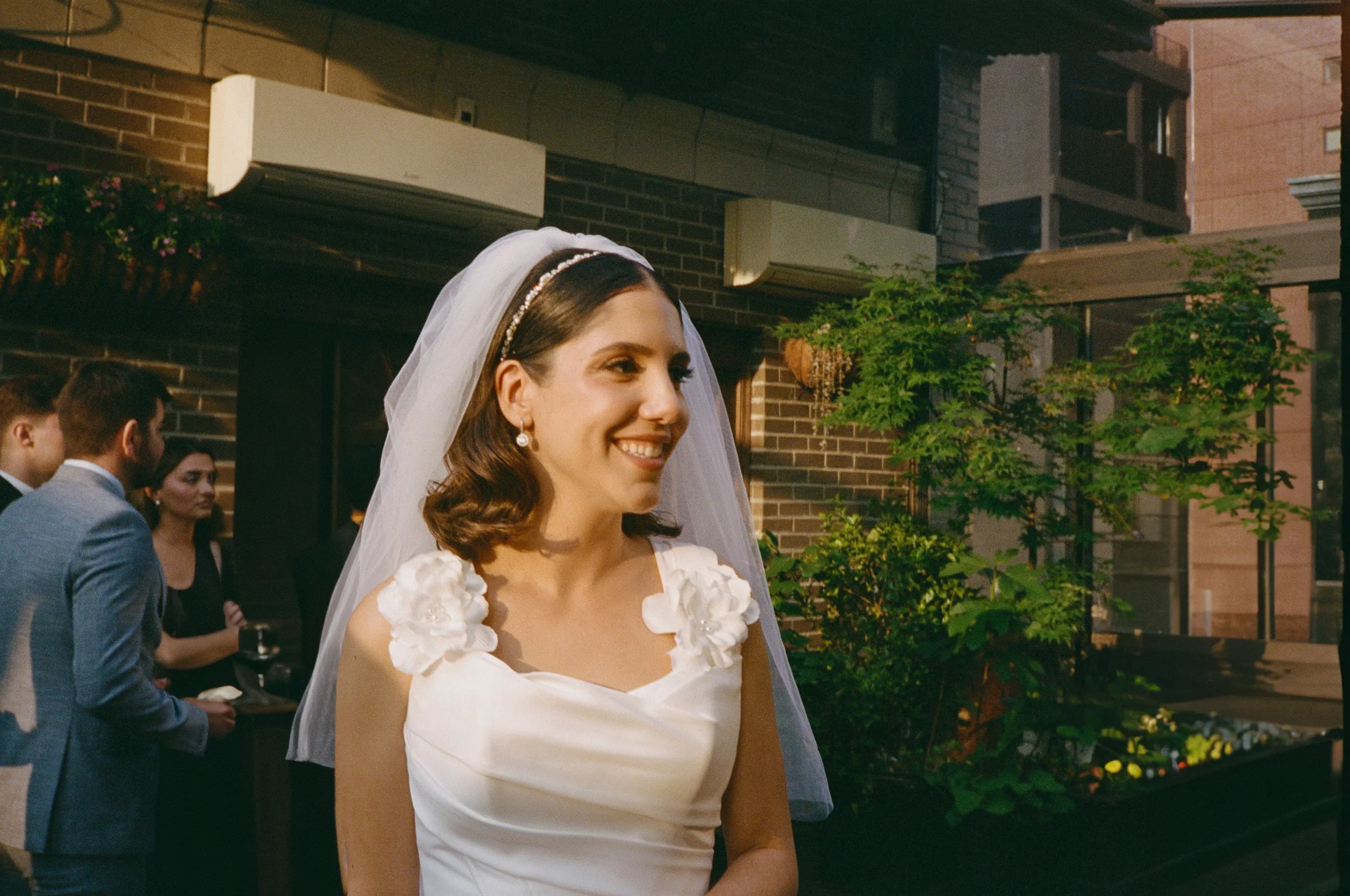 A bride with shoulder-length brown hair wearing a white wedding dress with floral shoulder details, a veil, and pearl earrings, smiles at a wedding reception outdoors.