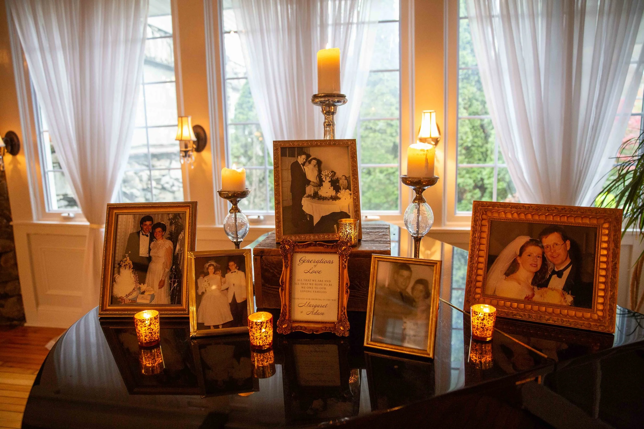 Display of framed wedding photographs, candles, and decorative items on a black table in front of a window with white curtains, inside a brightly lit room.