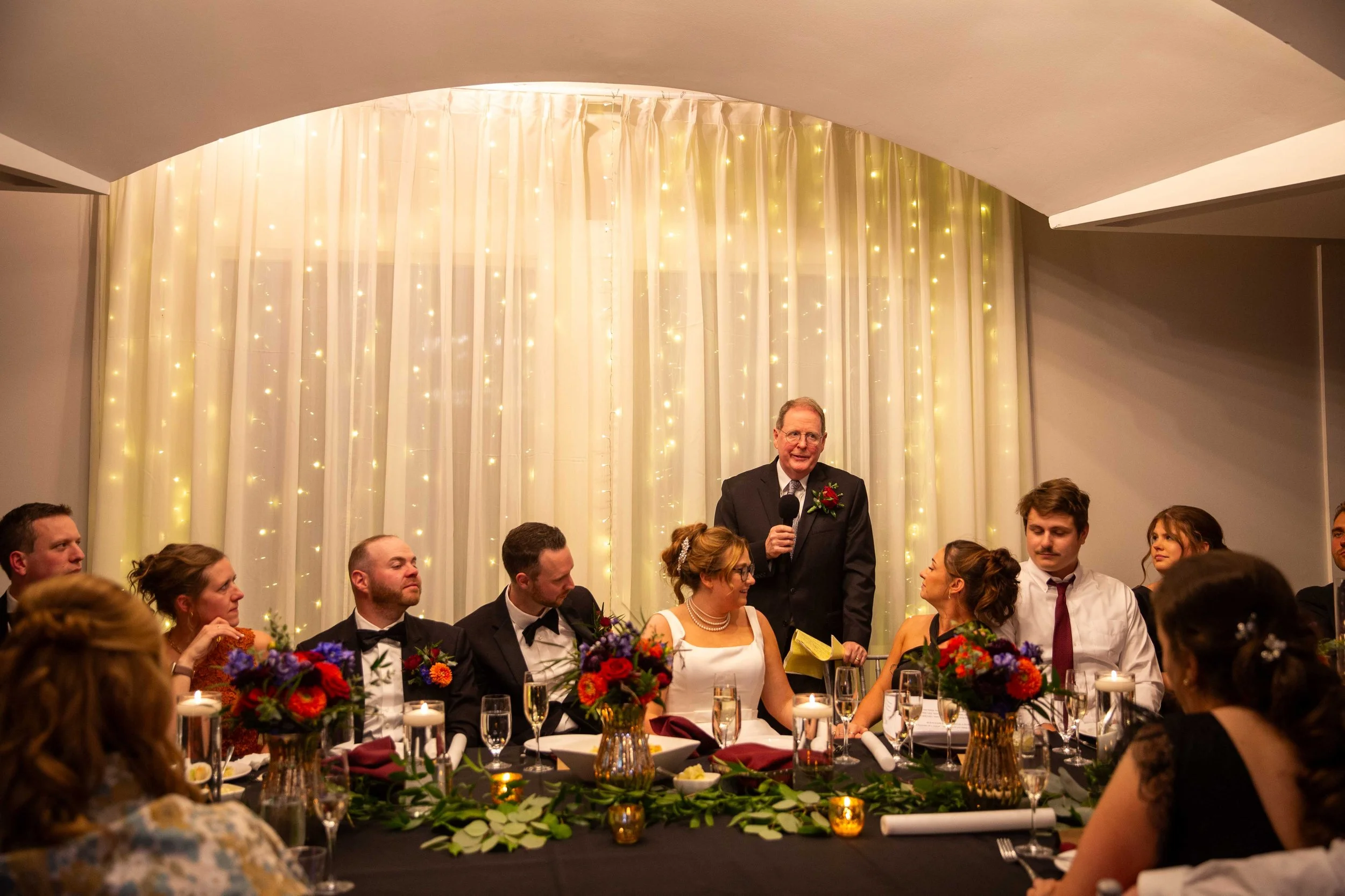 A wedding reception with a groom giving a speech, seated along a long banquet table with bridesmaids and groomsmen. The backdrop features curtains illuminated with fairy lights.