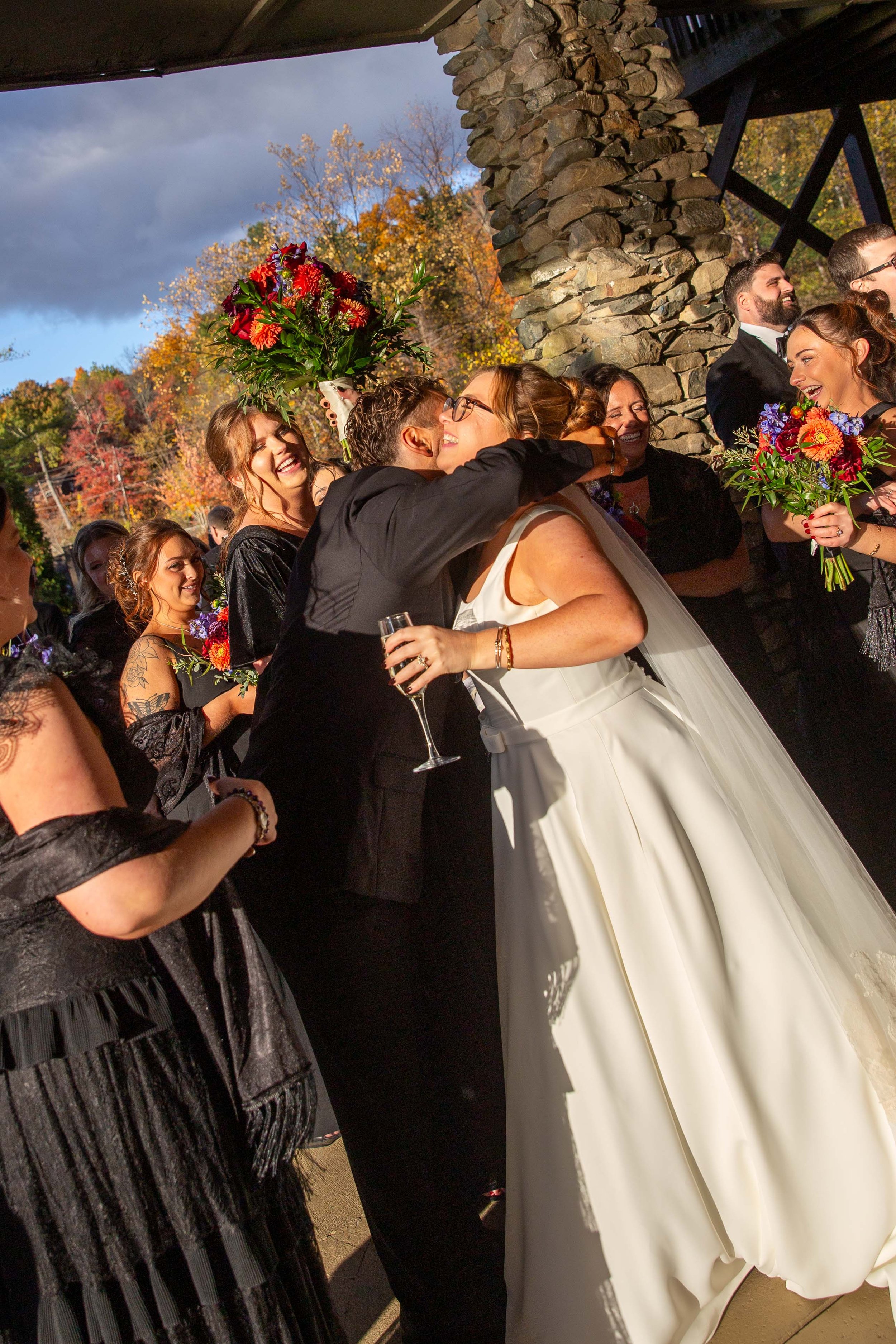 A group of people at a wedding celebration, with one person giving a hug to the bride who is holding a glass of champagne. The group includes women holding colorful bouquets, and they are standing under a stone and wooden structure with autumn trees 