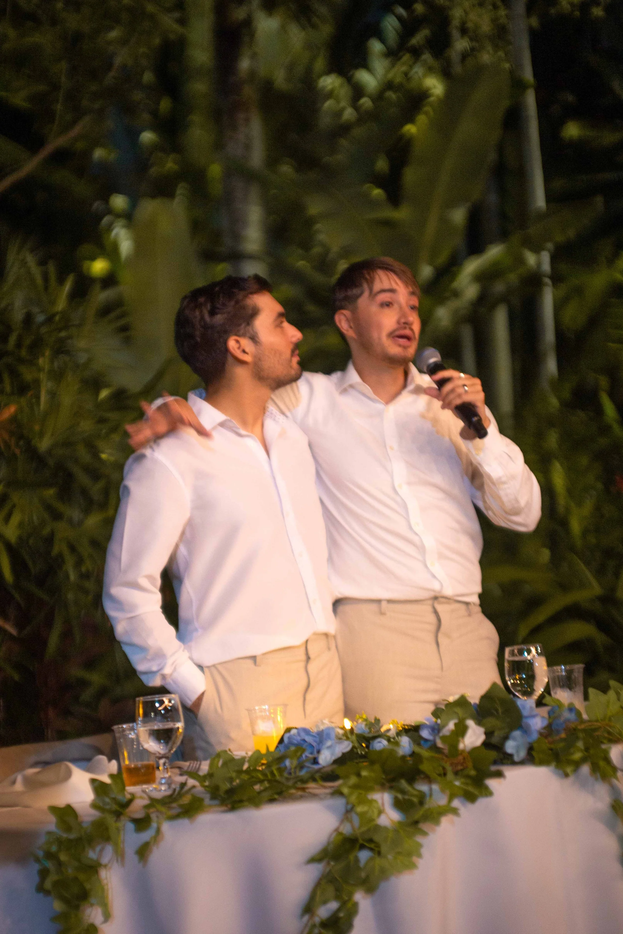 Two men in white shirts and beige pants stand together, one holding a microphone, with lush green foliage in the background, possibly at an outdoor event or celebration.