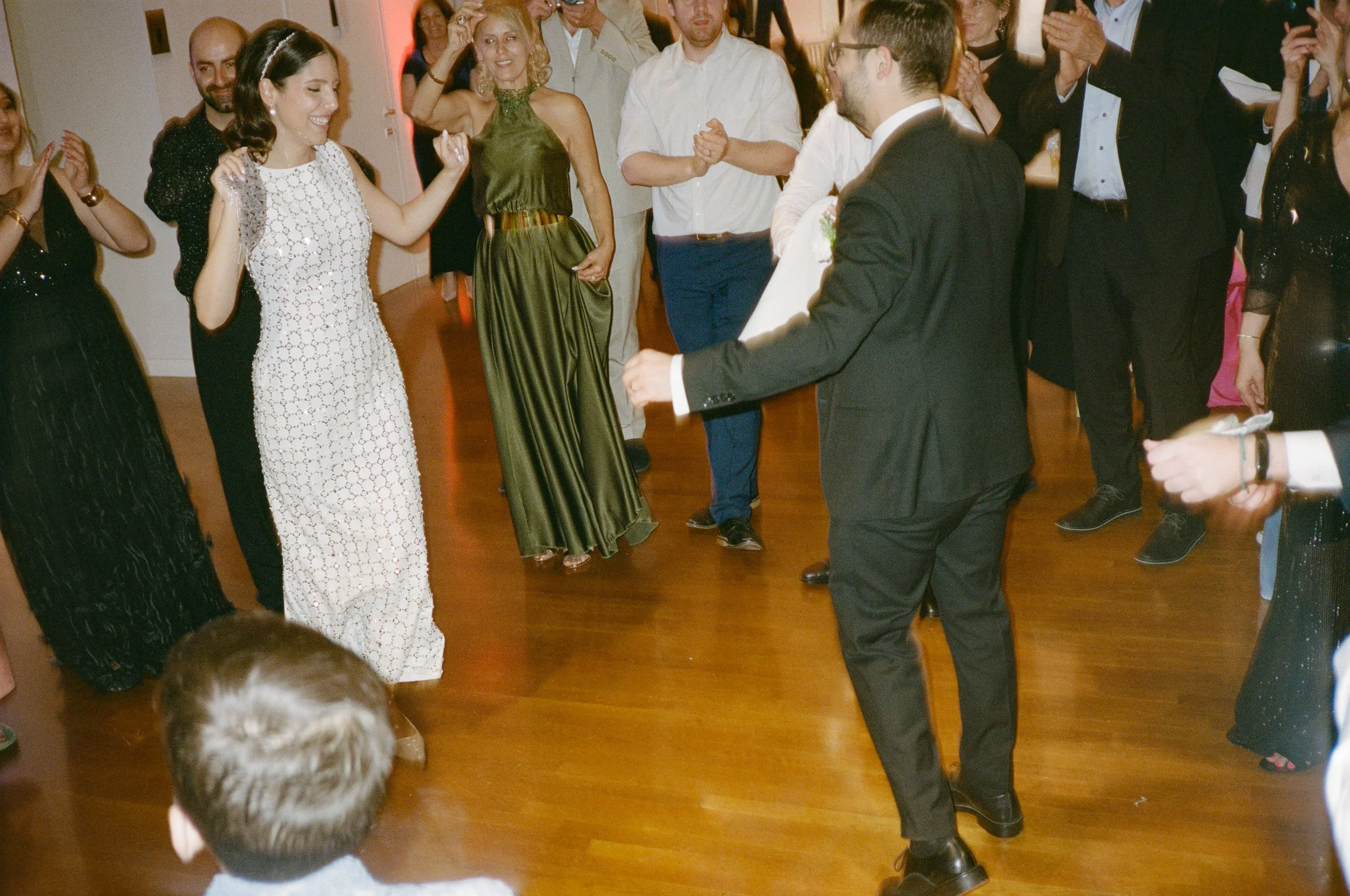 People dancing and celebrating, with a woman in a white dress and a man in a black suit in the center, at a formal event or wedding reception.