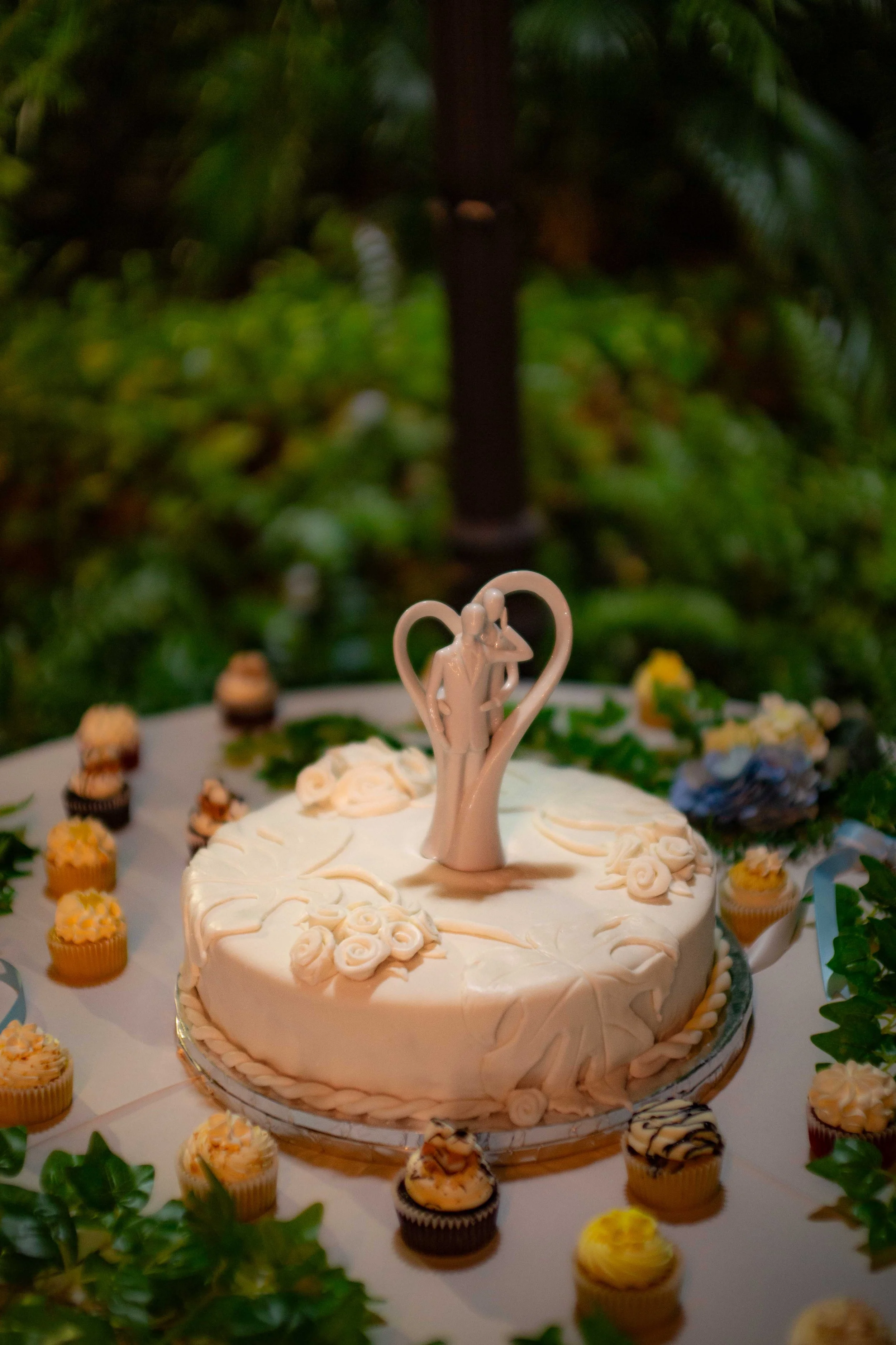 Wedding cake with a bride and groom topper, surrounded by cupcakes and greenery.