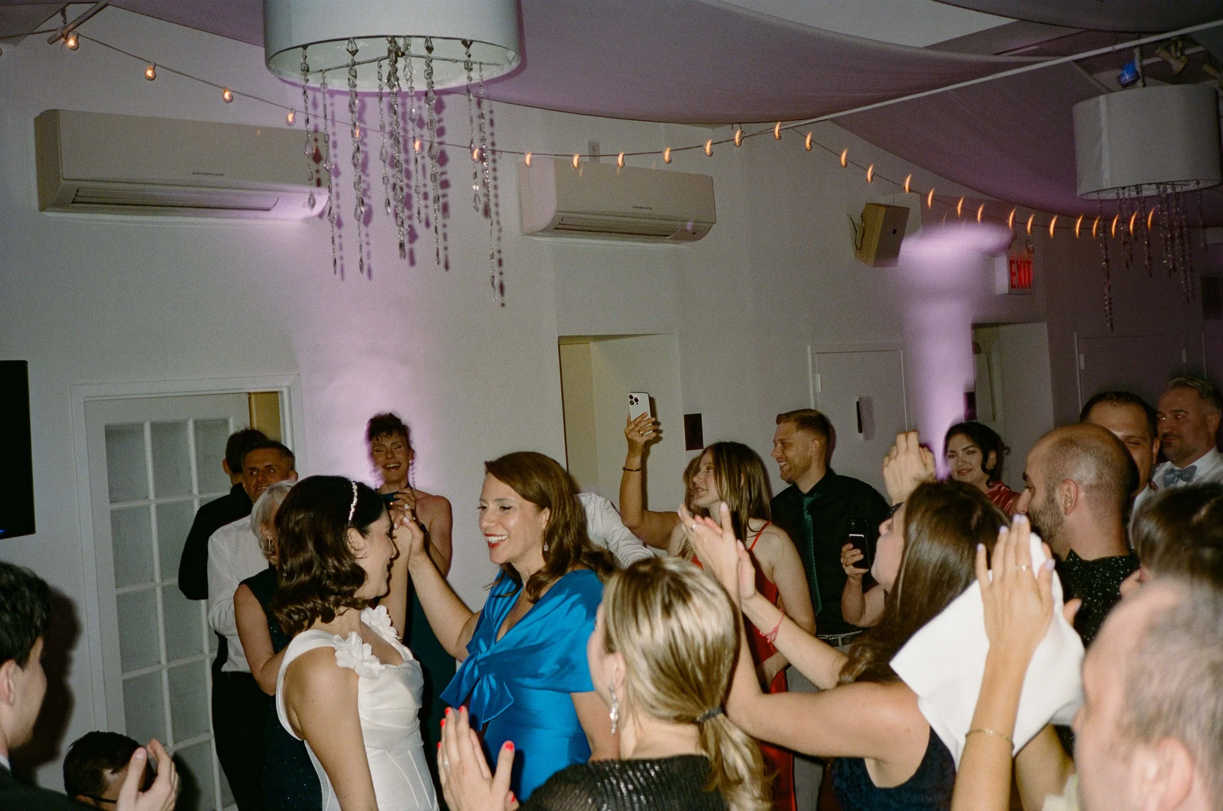 A group of people at a celebration, including a woman in a white dress, likely a wedding, smiling and interacting on a dance floor with others, some holding phones, in a decorated indoor venue with string lights and chandeliers.