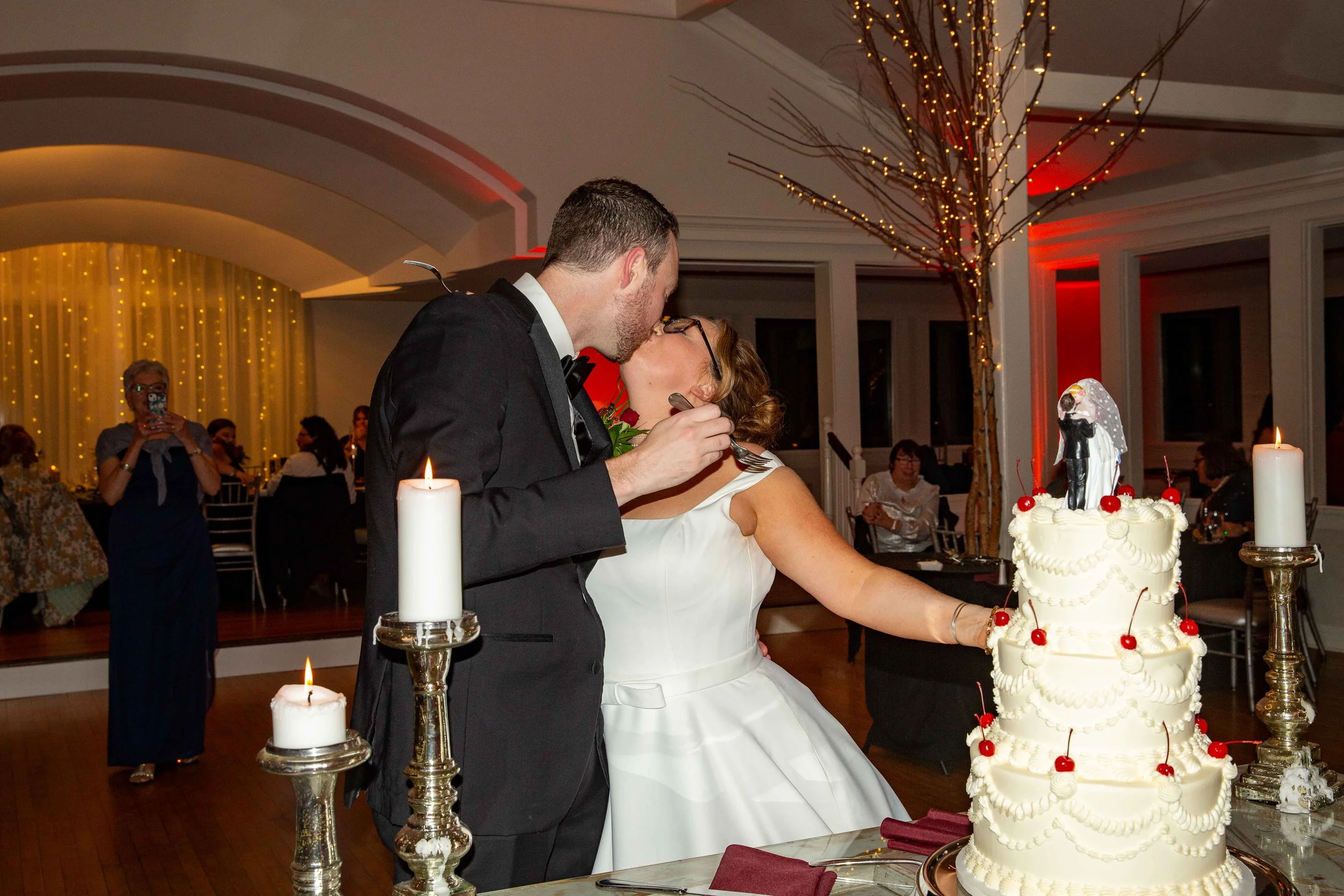A newlywed couple sharing a kiss at their wedding reception. The groom is in a black tuxedo and the bride is in a white wedding dress. They are standing in front of a multi-tiered wedding cake decorated with cherries and a bride and groom cake topper