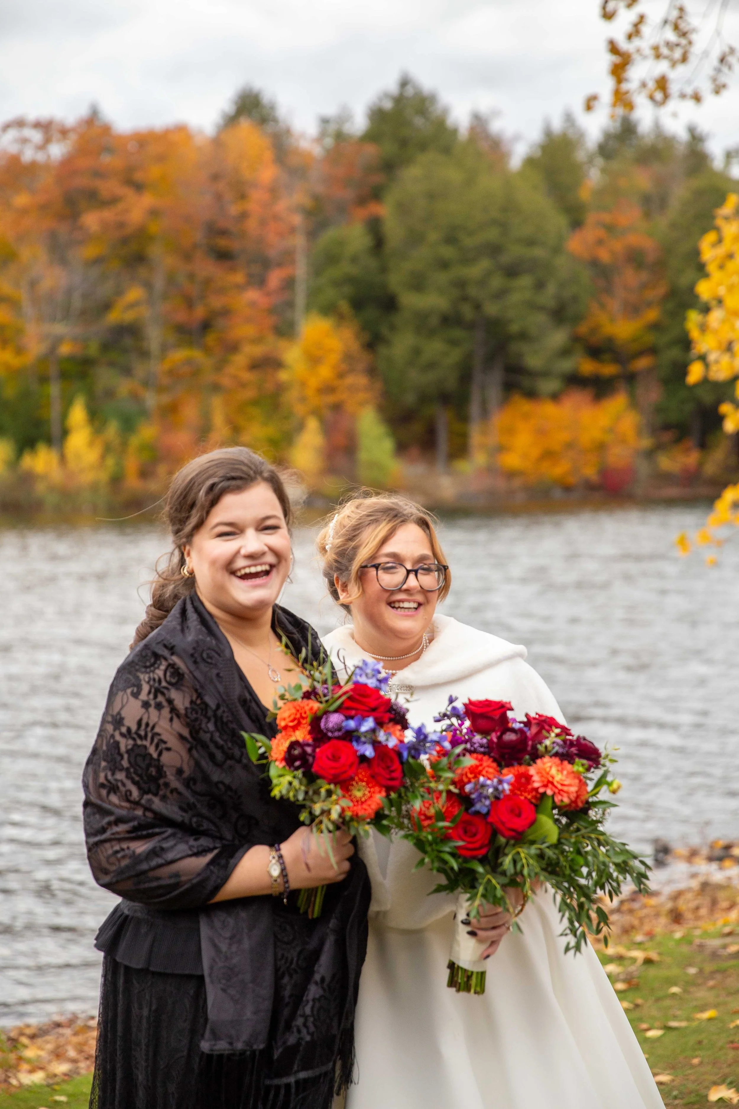 Two women smiling and holding large bouquets of colorful flowers by a lake with autumn trees in the background.