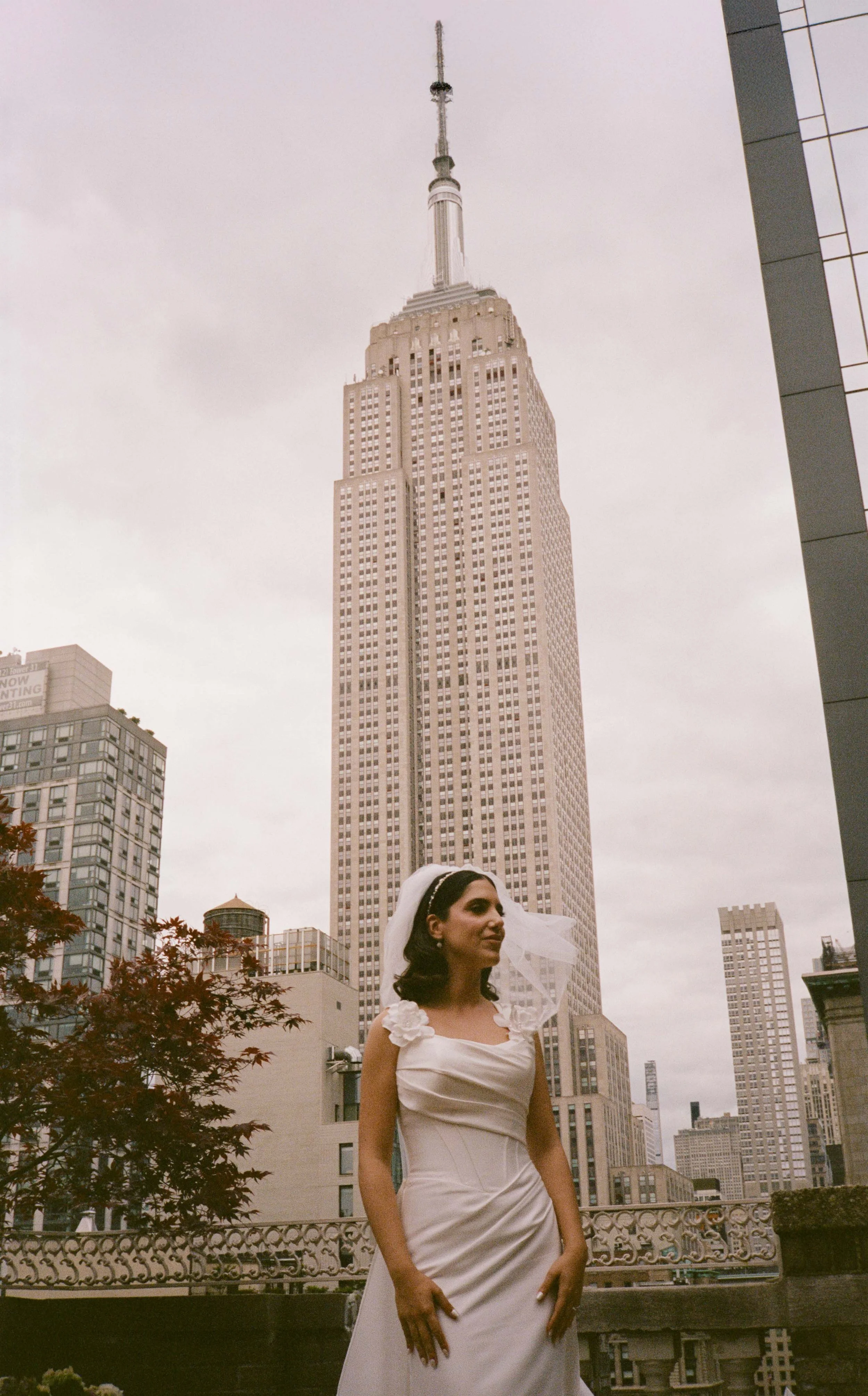 A woman in a wedding dress standing outdoors on a balcony with the Empire State Building in the background.