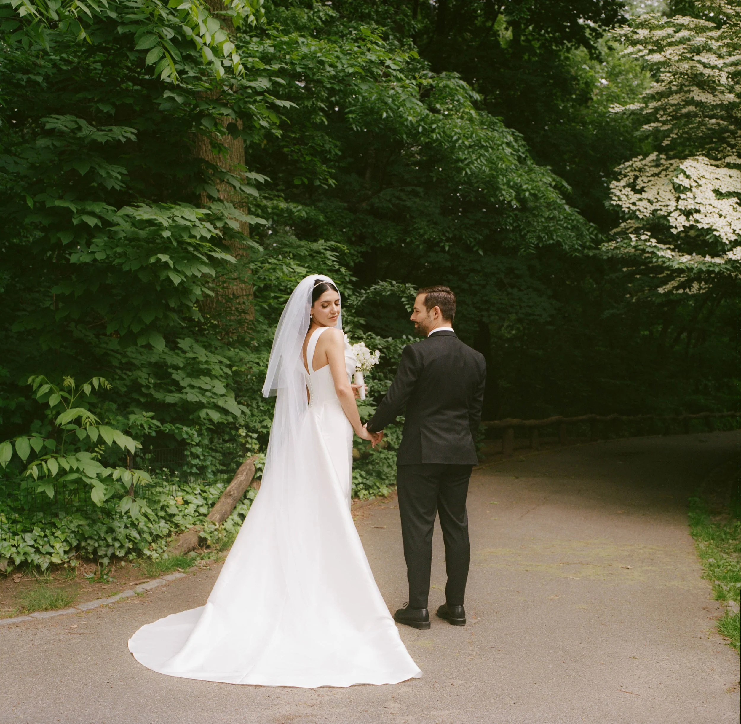 Bride and groom holding hands on a park pathway, surrounded by green trees and foliage, during their wedding.