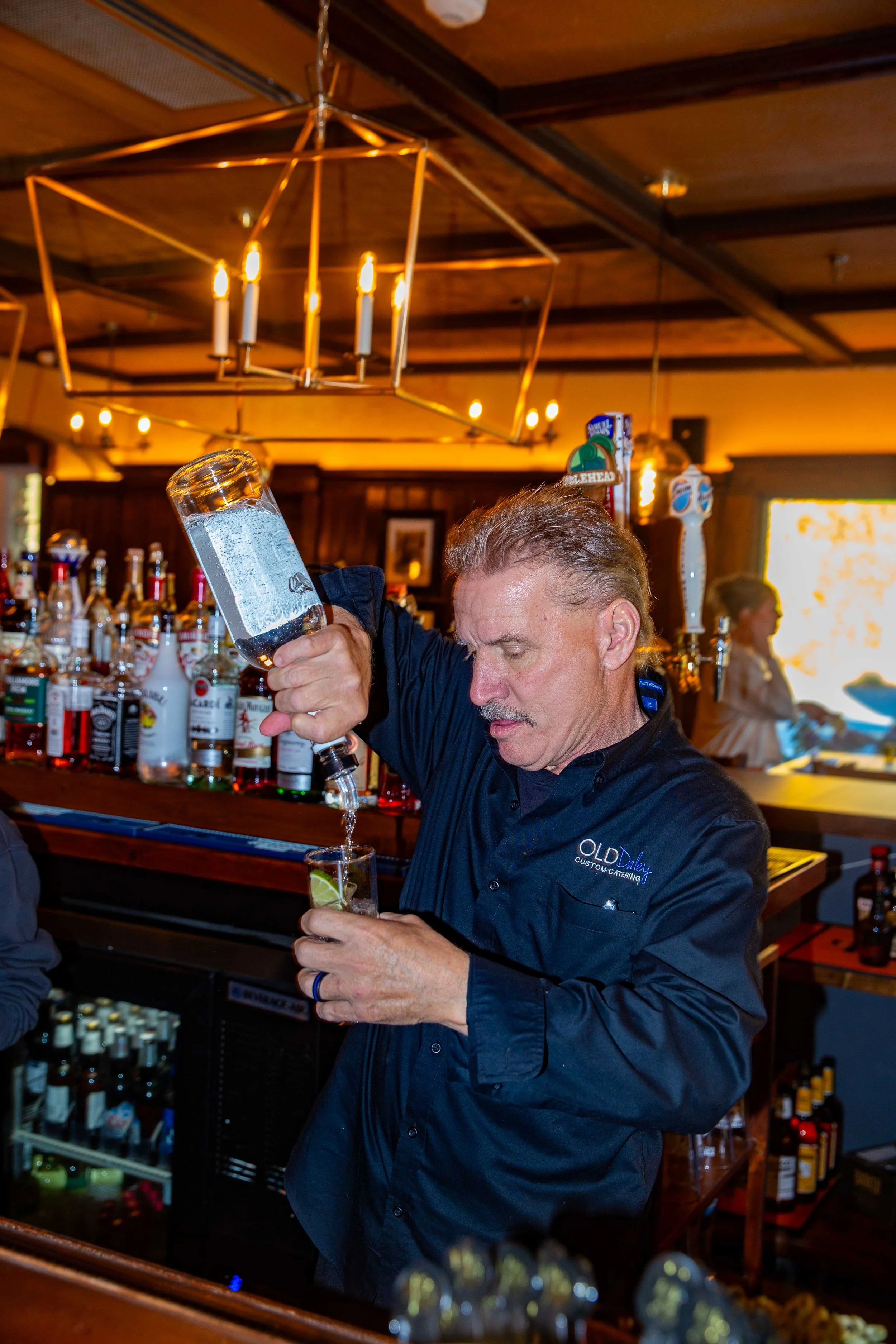 A bartender pouring a clear drink with lime into a glass at a bar, with bottles of alcohol on the shelves behind him.