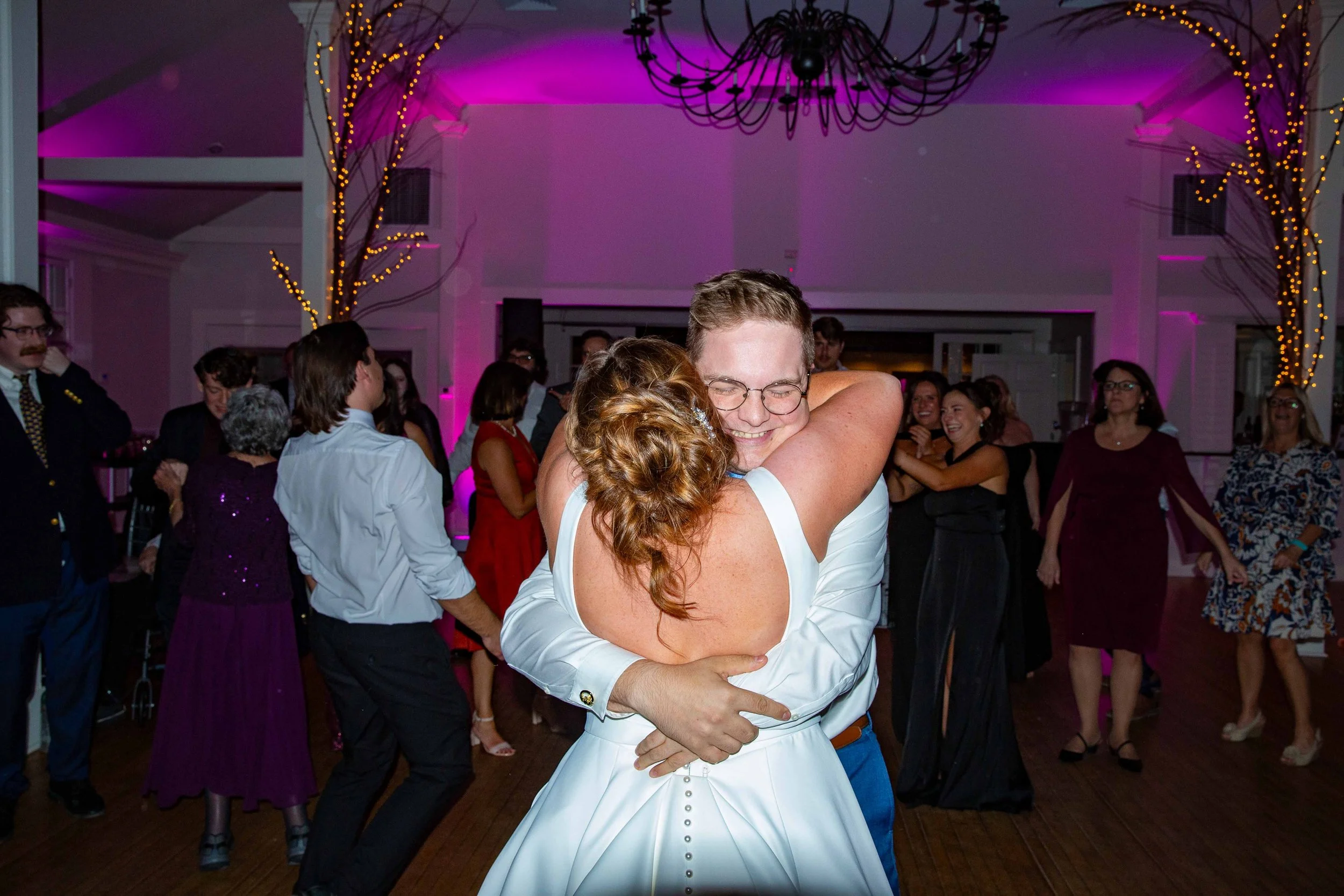 A couple dancing and hugging at a party, surrounded by other guests dancing and socializing in a decorated room with purple lighting and string lights.