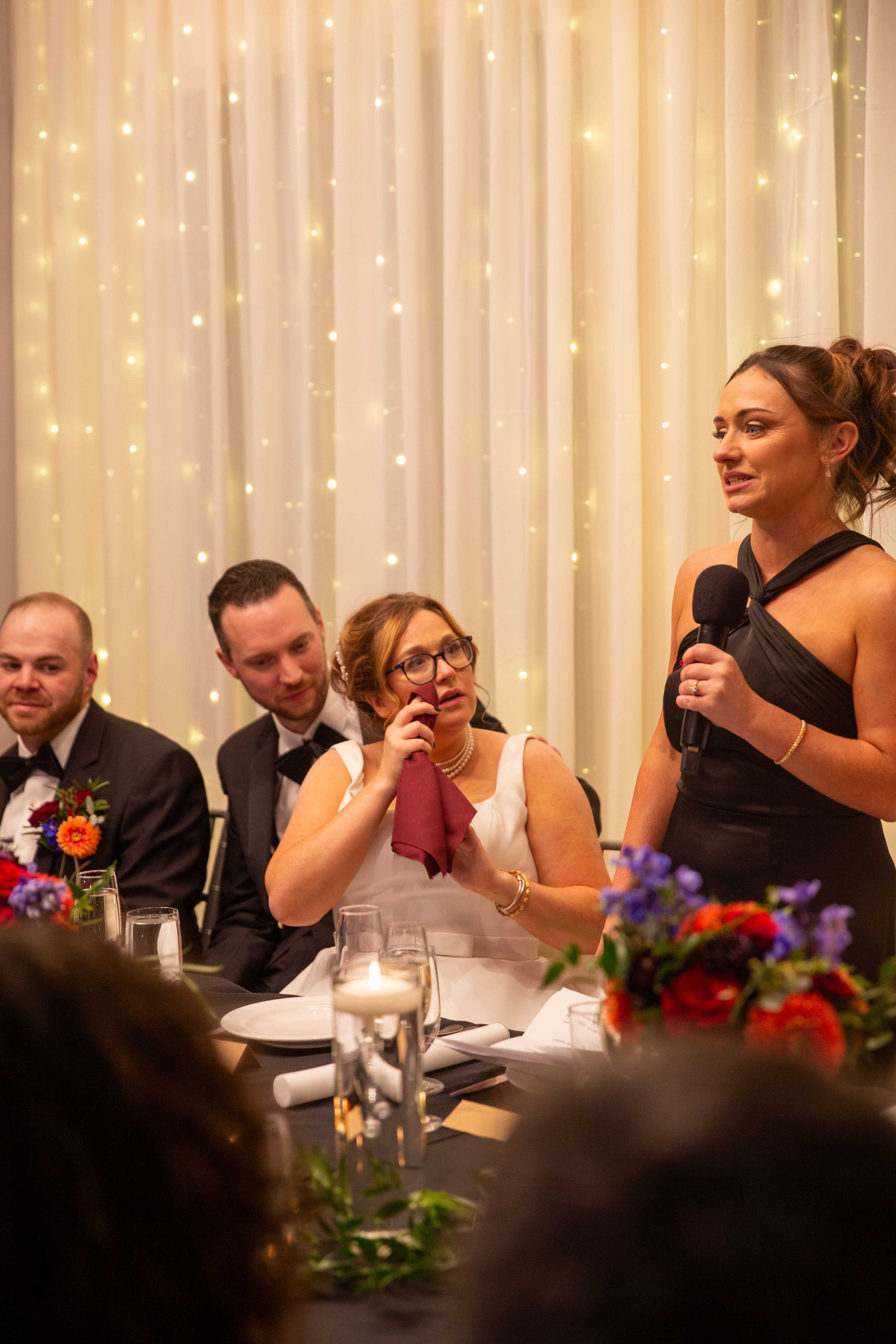 A woman in a black dress giving a speech at a wedding reception, with guests listening. The background has curtain with string lights, and the table has flowers and candles.